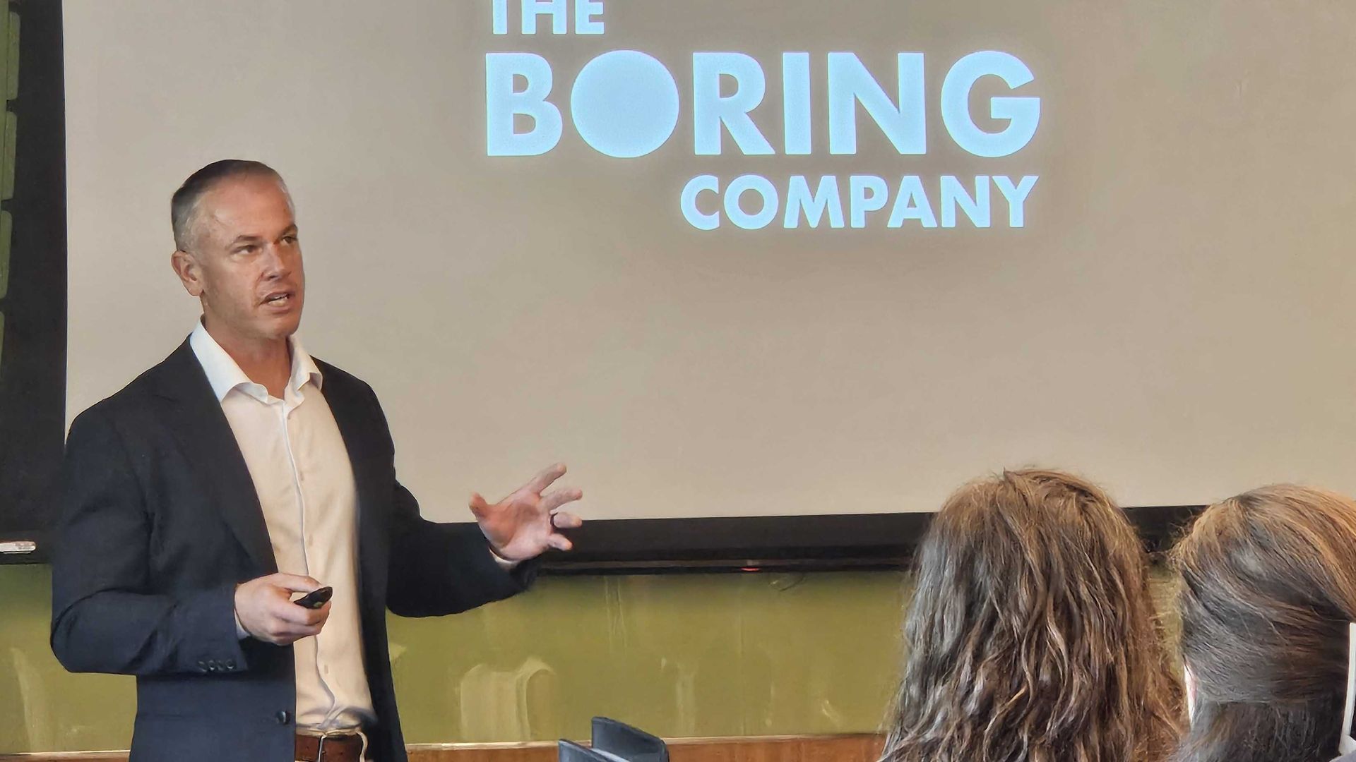Man in a suit giving a presentation with a screen showing "The Boring Company" logo while an audience watches.
