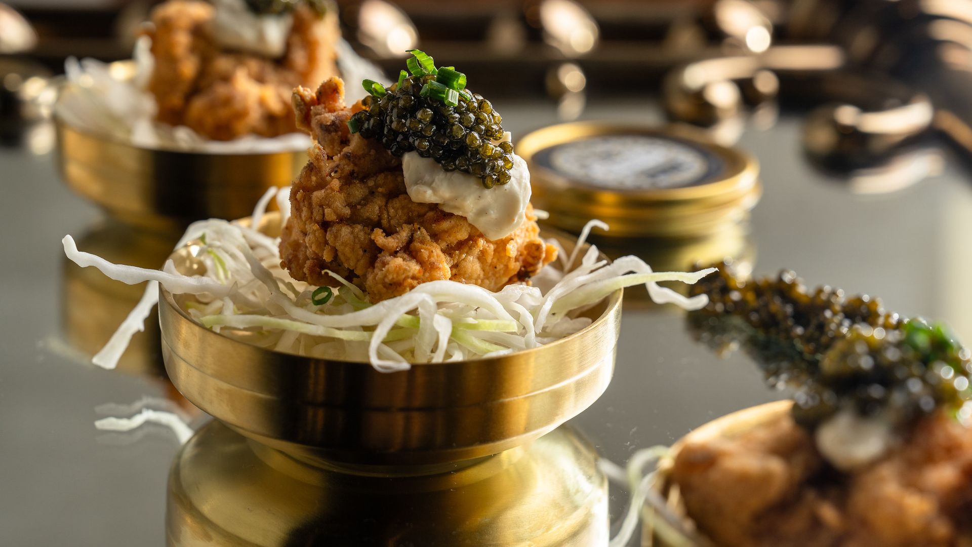 Close-up of a crispy fried bite topped with pale sauce, black caviar, and green onions, resting on shredded white cabbage in a gold serving cup; blurred background shows another similar dish.