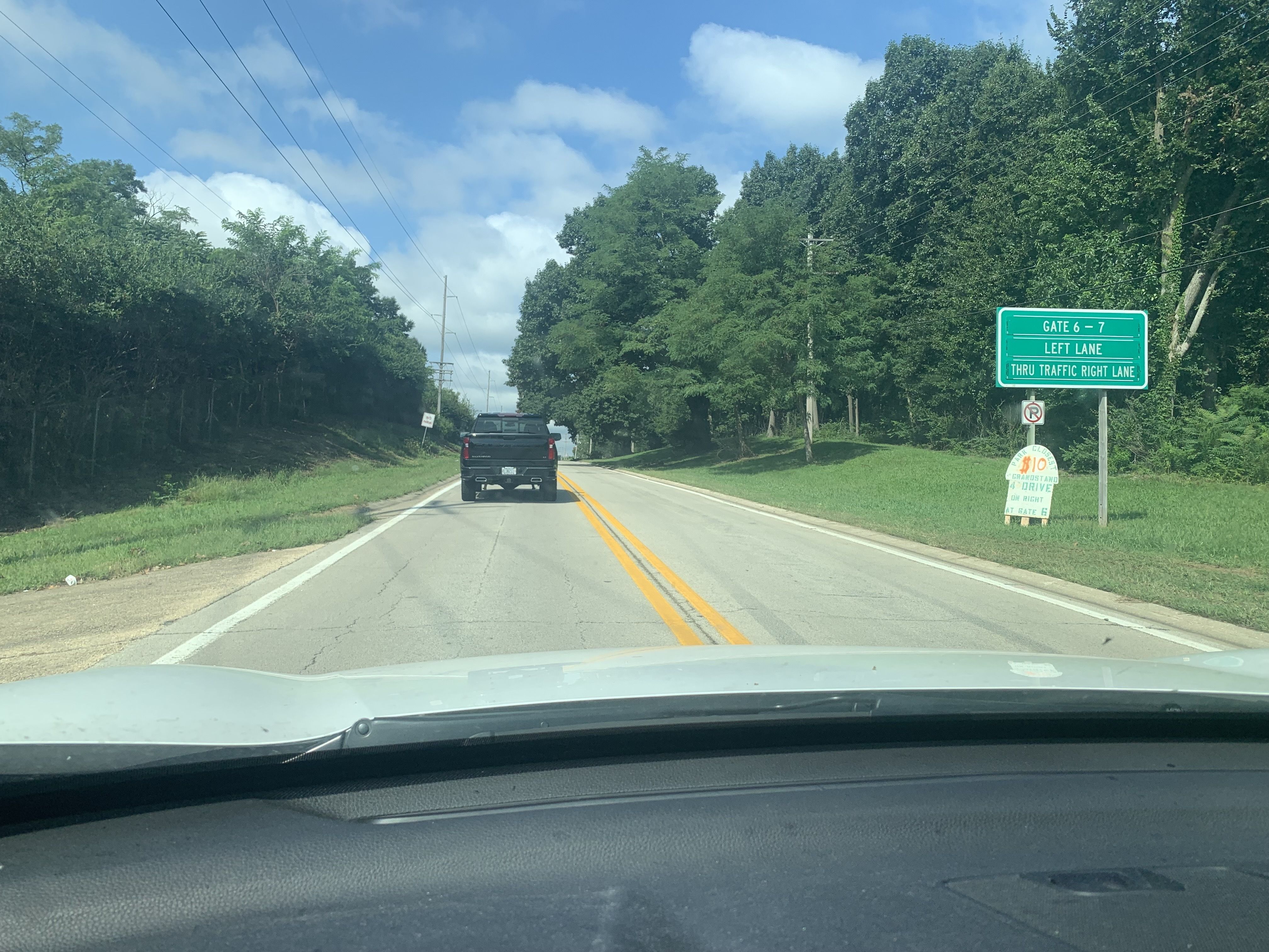 Two-lane road with dense green trees on both sides under a blue sky. A black pickup truck follows. A green highway sign on the right reads, "GATE 6-7 LEFT LANE THRU TRAFFIC RIGHT LANE."