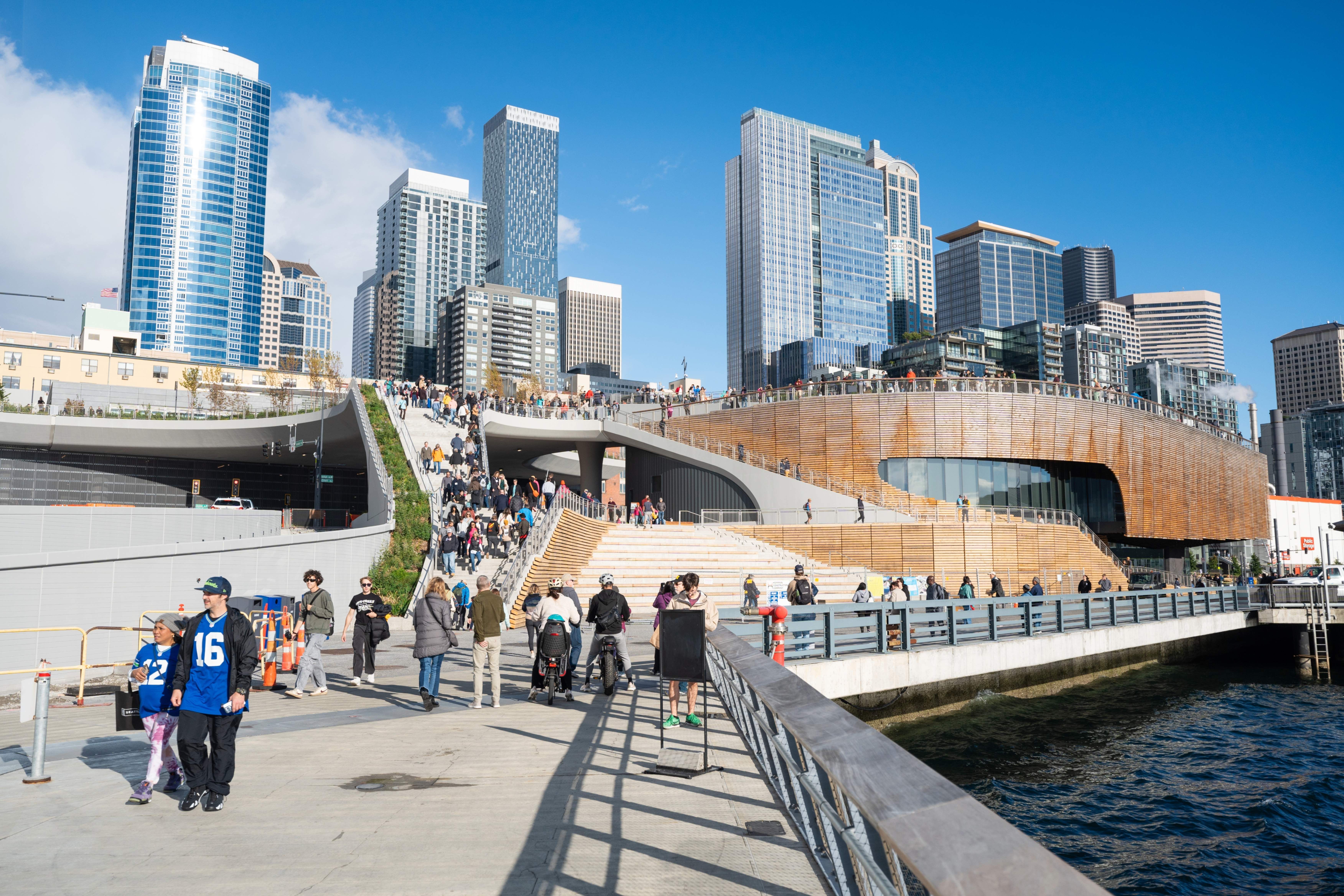 People walk by the Overlook Walk and the Seattle Aquarium's new Ocean Pavilion on the Seattle waterfront, with the Seattle skyline in the background.