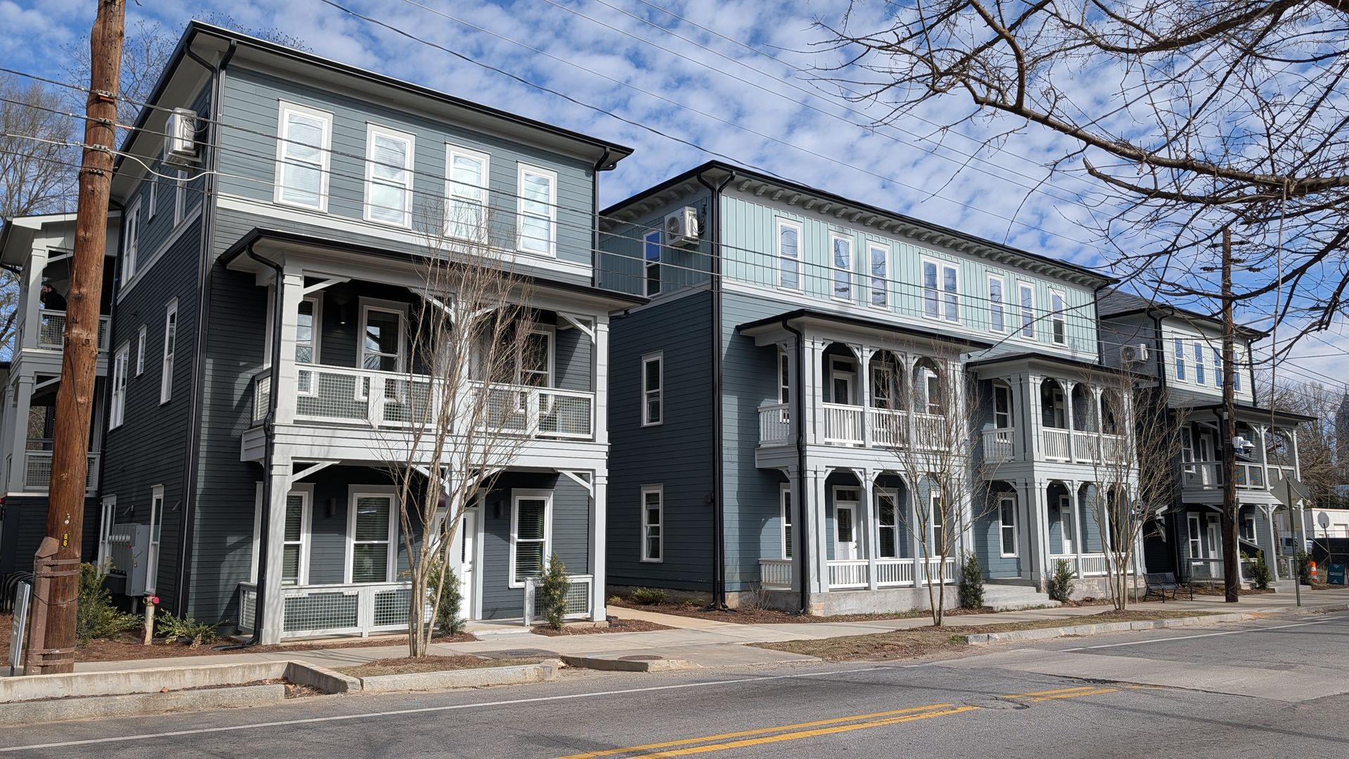 Three modern blue-gray apartment buildings with white trim and balconies along a street lined with leafless trees under a partly cloudy blue sky.