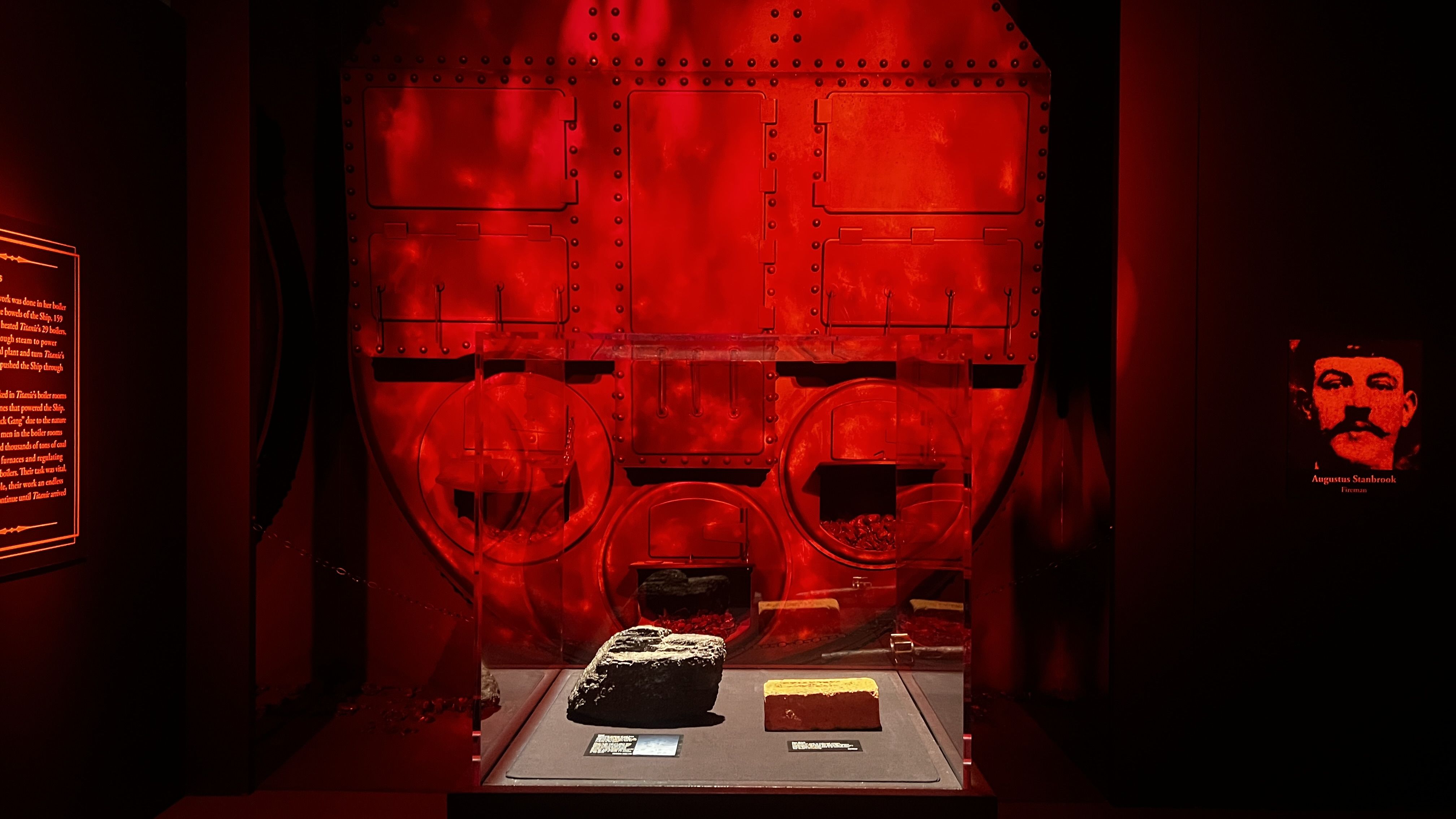 Red-lit museum exhibit of a large riveted metal boiler with rectangular doors, rivets, and pipes; two circular wheels at the bottom. In front are rocks on a pedestal, with left text panel and a right portrait plaque of a man.