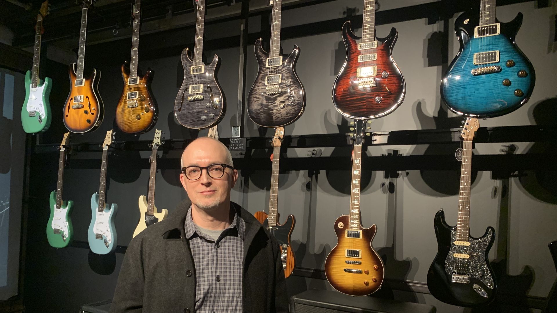 Man with glasses and a gray coat stands in front of a wall displaying various electric guitars in green, blue, black, brown, and sunburst finishes.