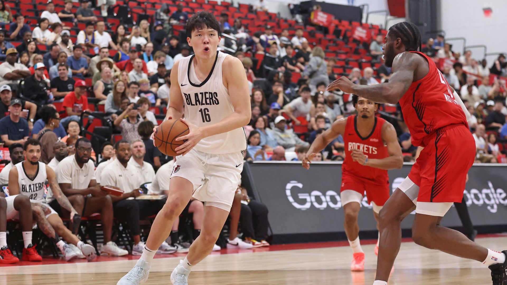 Basketball player Yang Hansen in white Blazers jersey #16 dribbling while guarded by two players in red Rockets jerseys during a game with spectators in the background.