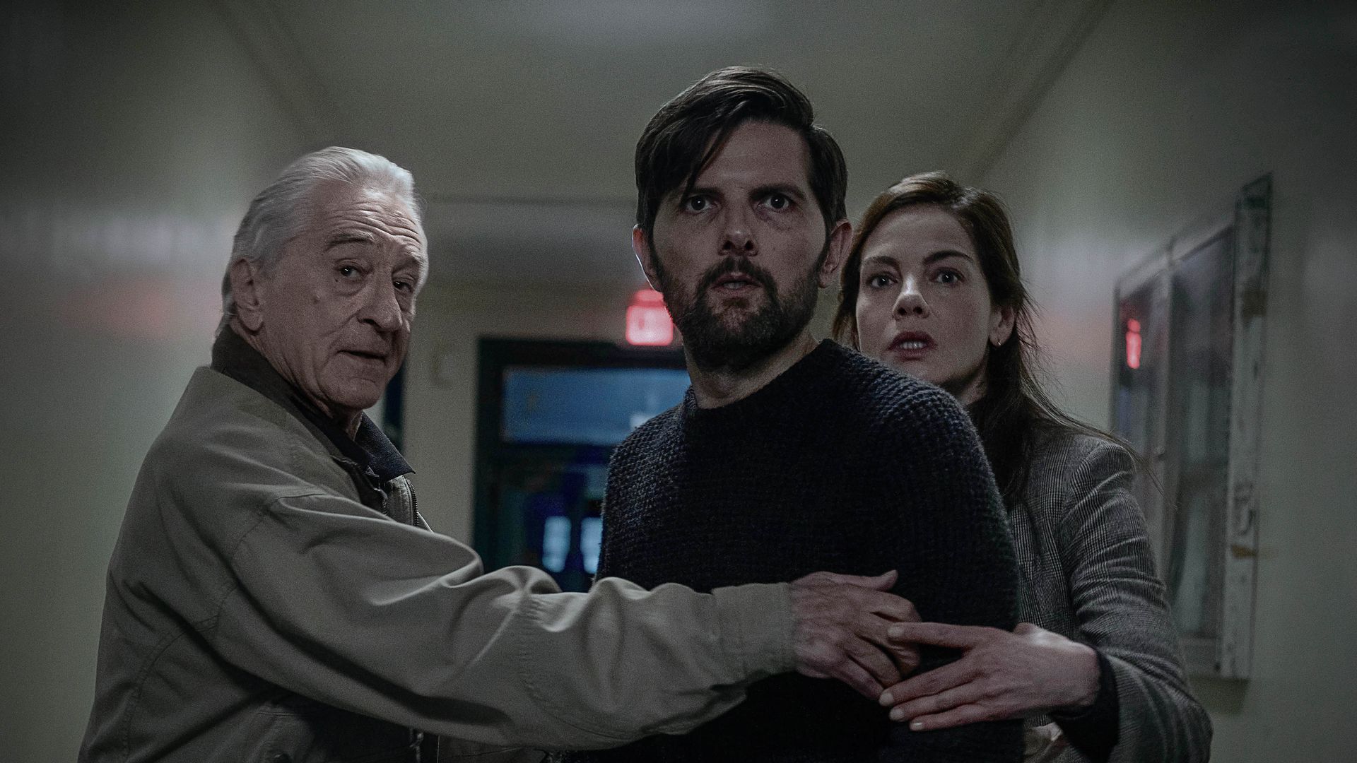 Three adults in a dim hallway, an older man on the left (Robert De Niro) embraces a bearded man in a dark sweater (Adam Scott) while a woman (Michelle Monaghan) clings behind them; tense, wary expressions with an exit sign in the background.