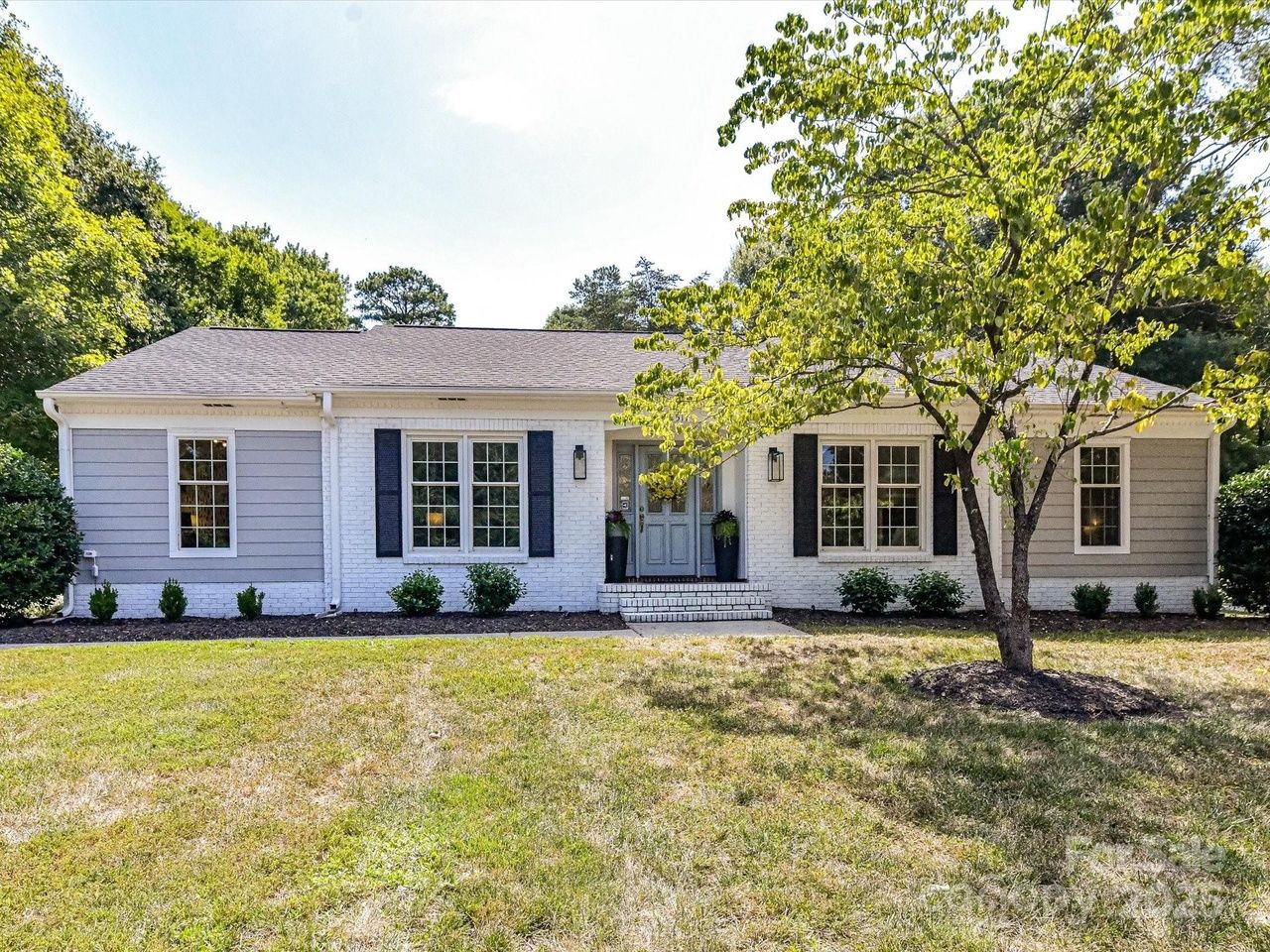 Single-story house with gray and white exterior, black shutters, a front door with sidelights, surrounded by green bushes and a tree on a lawn under a sunny sky.