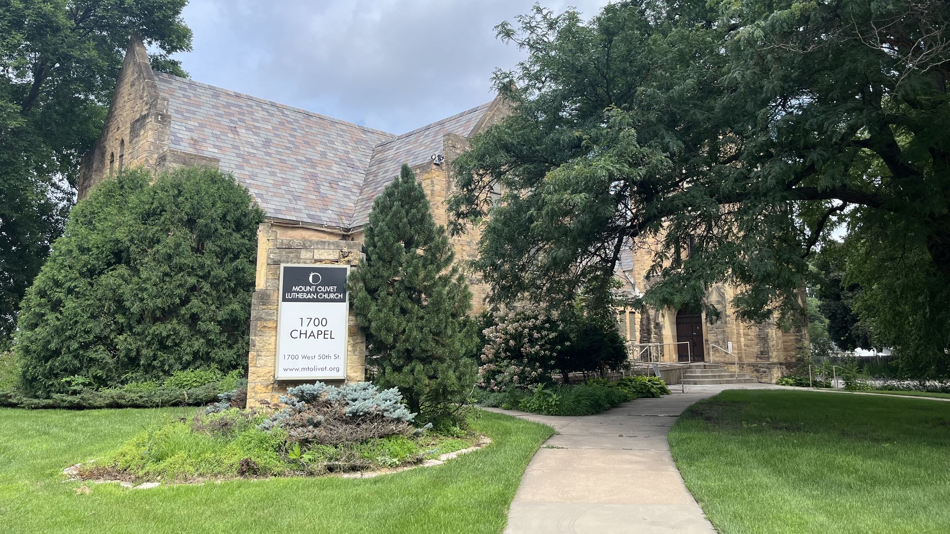 Stone chapel building with slate roof, surrounded by green trees and grass. Sign reads "Mount Olivet Lutheran Church 1700 Chapel 1700 West 50th St." under cloudy sky.