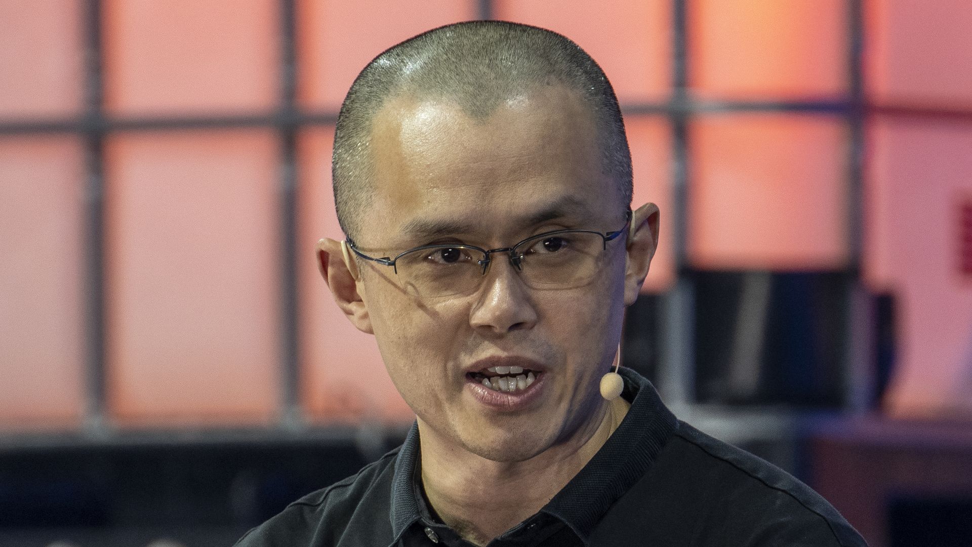 Changpeng Zhao looking toward and the left of the camera, speaking to an audience it a small microphone extending from his ear.