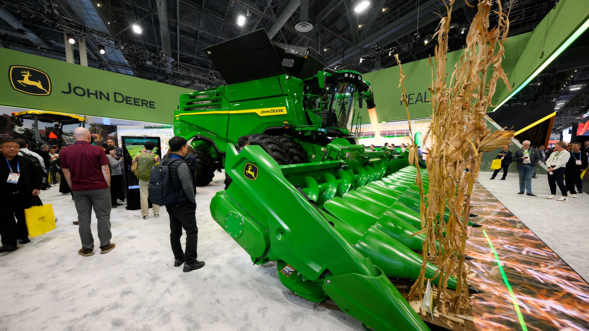 Green John Deere combine harvester displayed indoors at an exhibition with people around, featuring large green harvesting equipment and cornstalks on the side.