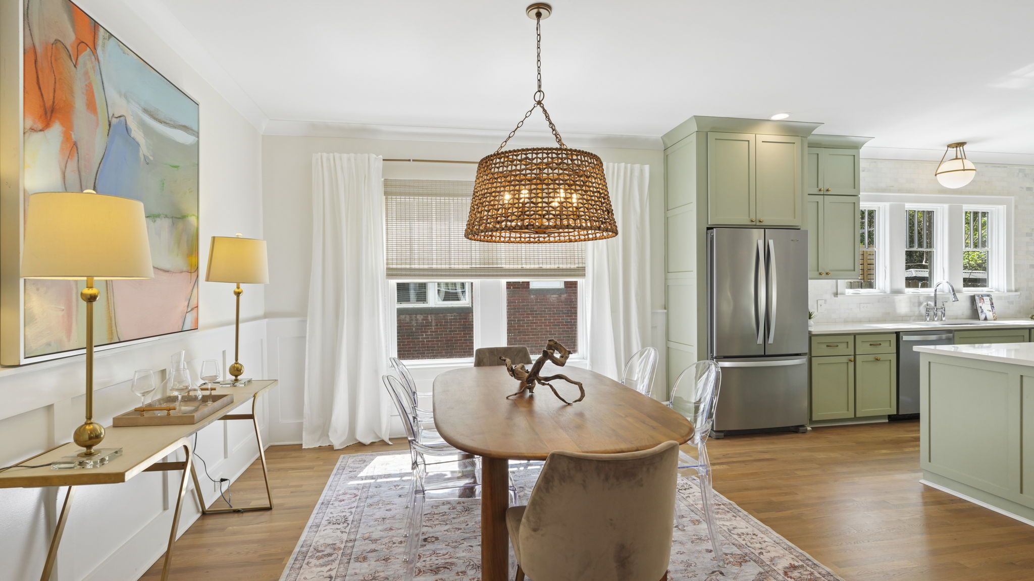 Bright dining room with oval wooden table and clear acrylic chairs under a woven rattan pendant light. Light wood floors, pale green cabinets, stainless fridge, white curtains, and abstract art.