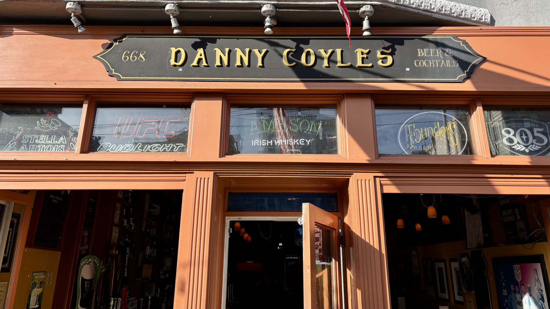 Front of bar named Danny Coyles with orange trim, black and gold sign reading "Danny Coyles Beer & Cocktails," and neon signs for Stella Artois, UFC Bud Light, Jameson Irish Whiskey, Founders, and 805.