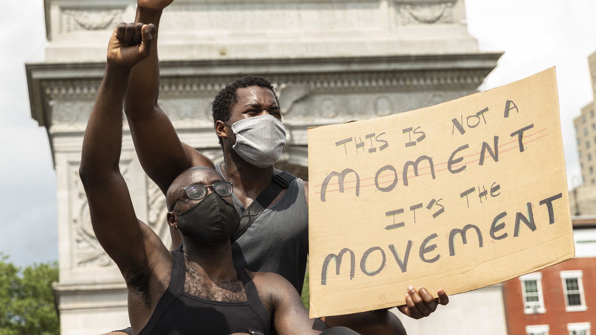 Photo of two Black protesters raising their fists in the air as one holds a sign that says "This is not a moment. It's the movement."