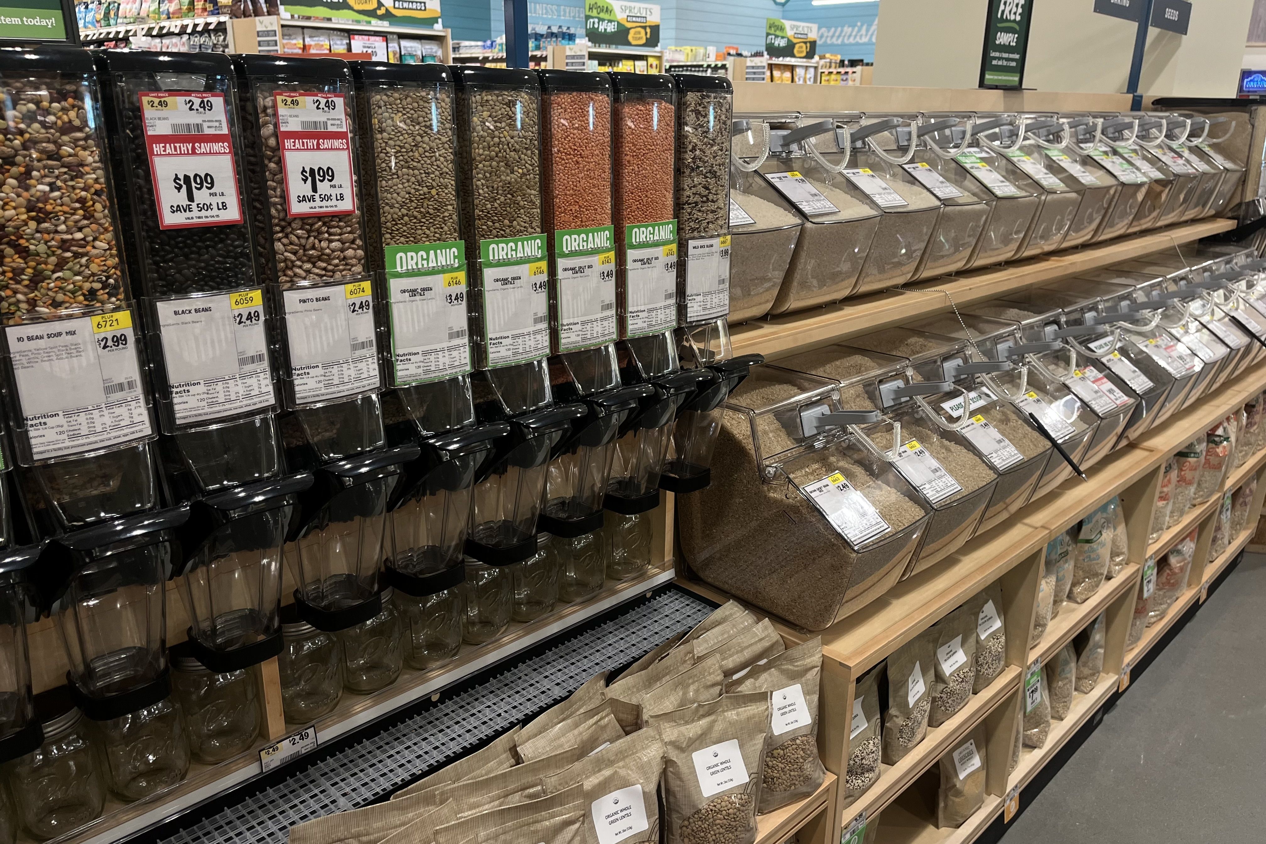 Grocery store bulk bins with various beans, lentils, and grains in clear containers labeled with prices and organic tags on wooden shelves.