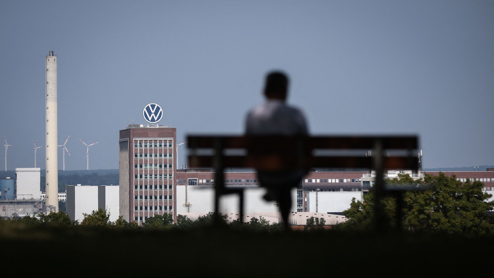 A person sits on a bench with an automotive factory in the background