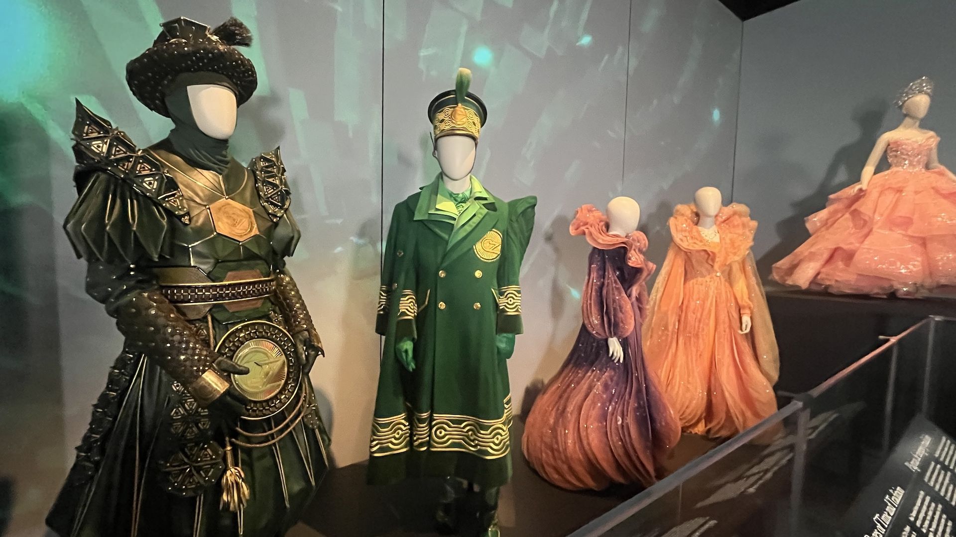 Display of five theatrical costumes on mannequins including a green armored suit with spikes, a green military-inspired coat with gold details, two elaborate purple and orange gowns, and a peach ball gown with a tiara.