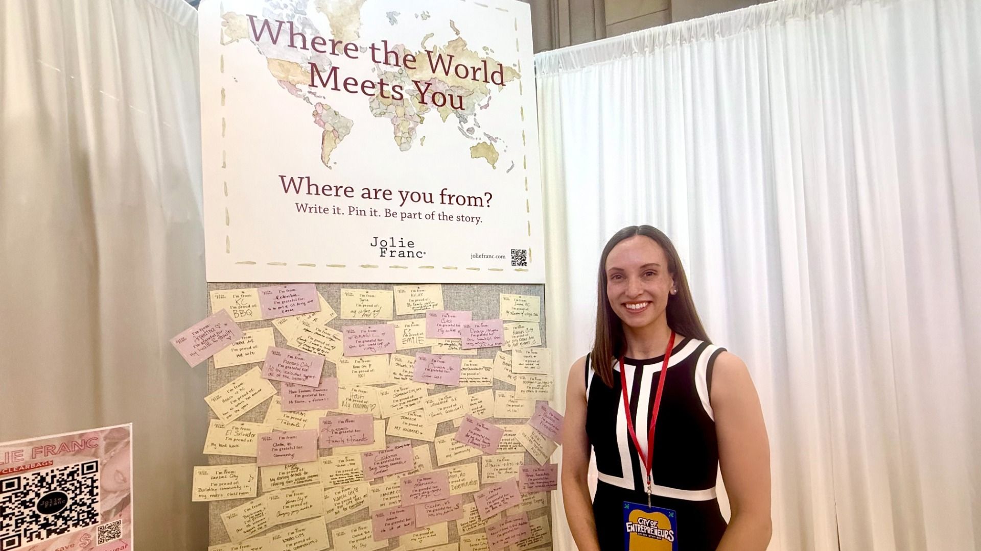 Smiling woman in a black-and-white dress stands beside a poster that says "Where the World Meets You" with a world map. Wall covered in pink and beige sticky notes; she wears a red lanyard.