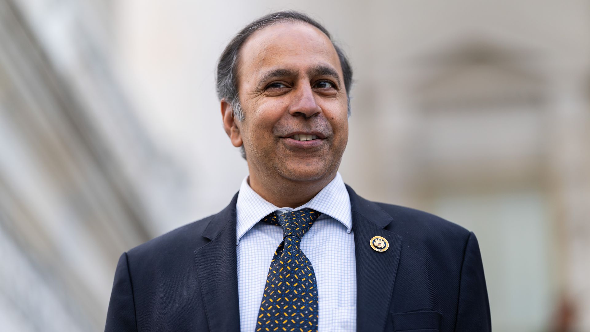 Close-up portrait of a smiling man in a dark suit, light blue checkered shirt, and a navy tie with yellow shapes. A round lapel pin is on his blazer; blurred stone background.