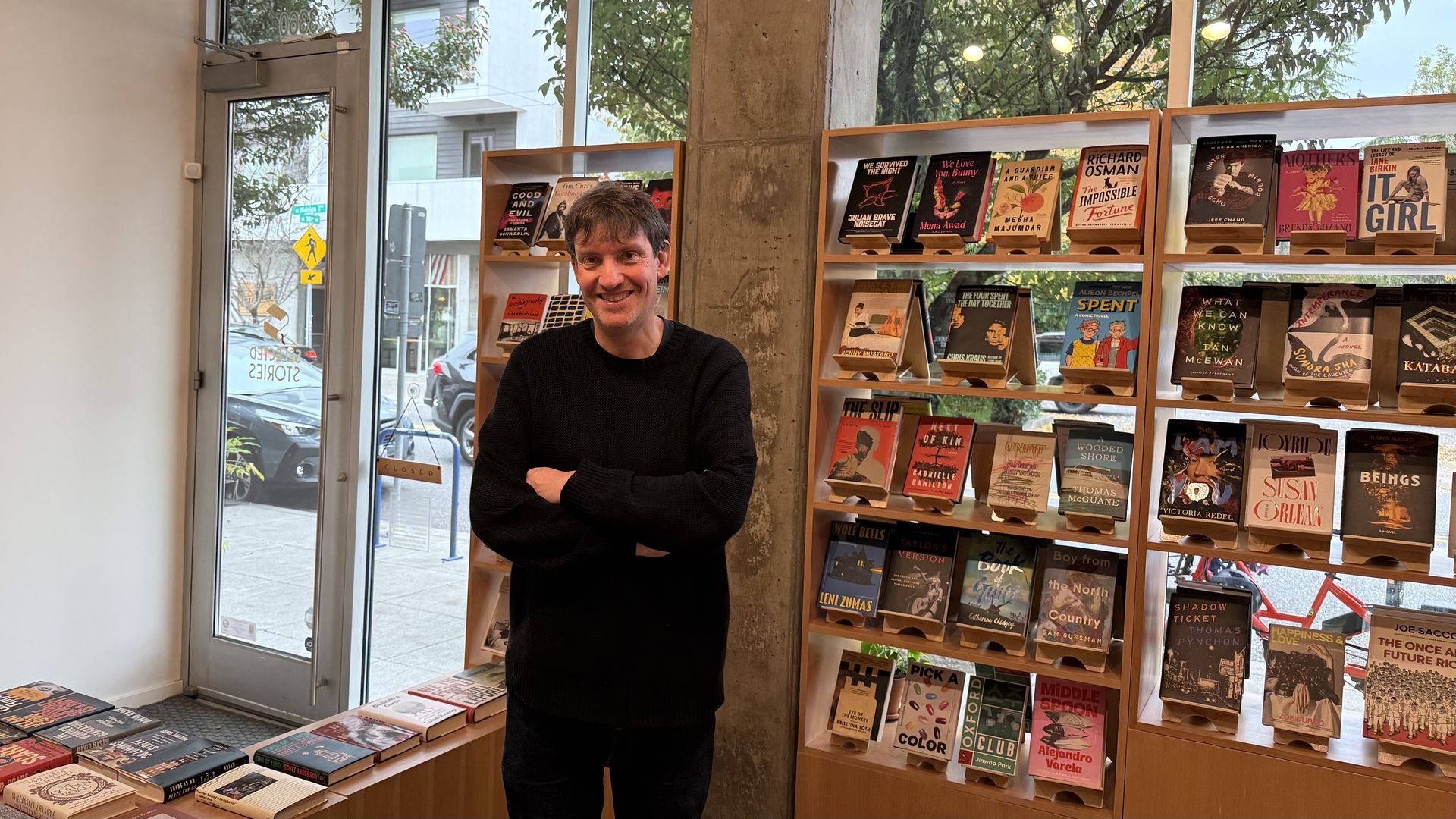 A person in a black sweater stands smiling inside a bookstore with wooden shelves filled with colorful books. Outside, cars and trees are visible through large windows and a glass door.
