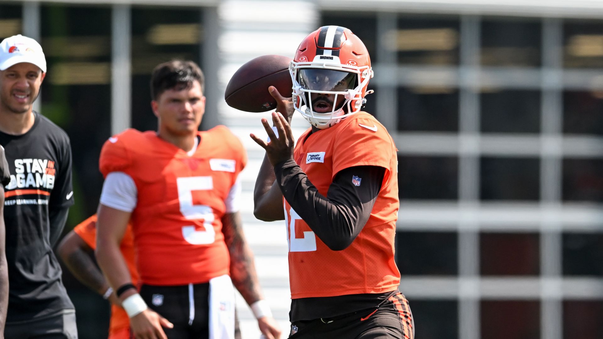 Cleveland Browns football player in orange jersey and helmet prepares to throw a football during practice, with teammates and coach in background near a building with large windows.