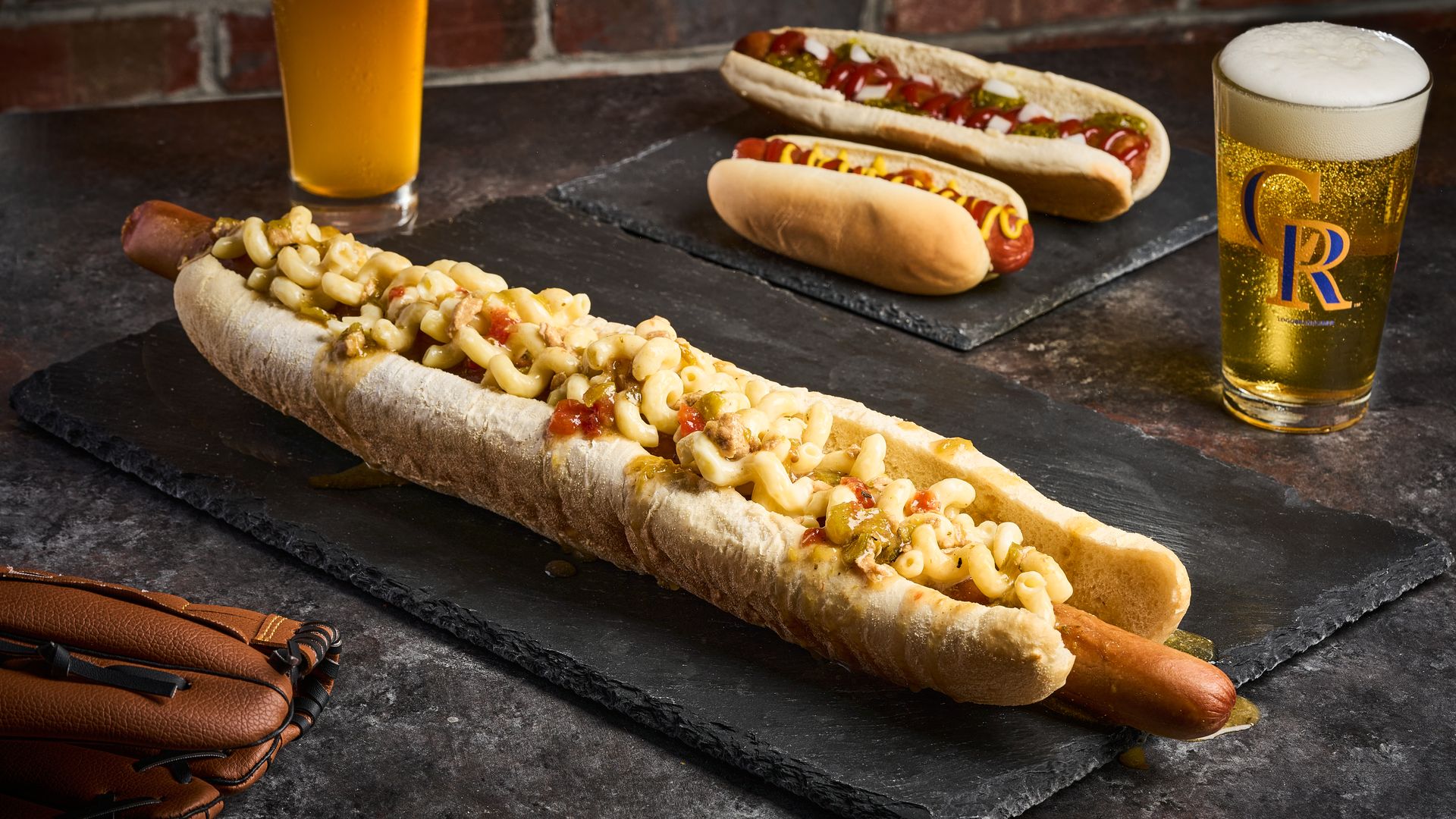 Long hot dog in a crusty bun topped with macaroni and cheese, with two other topped hot dogs on a slate board in the background; beer glasses and a leather glove nearby.