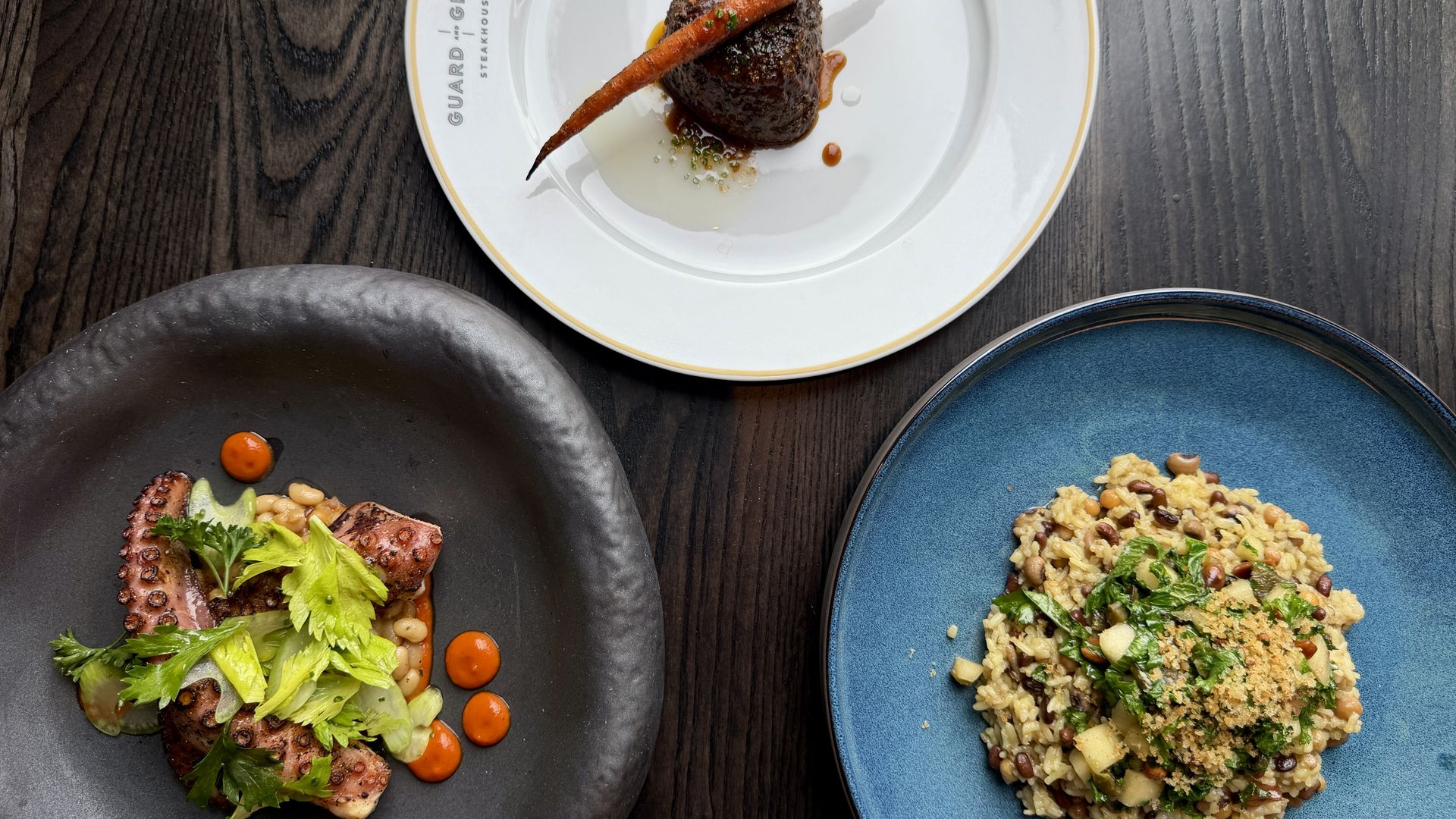 Top-down view of a dark wooden table with three plates: a white plate holding a seared steak and a long carrot, a black plate with octopus and greens, and a blue plate of risotto with beans.
