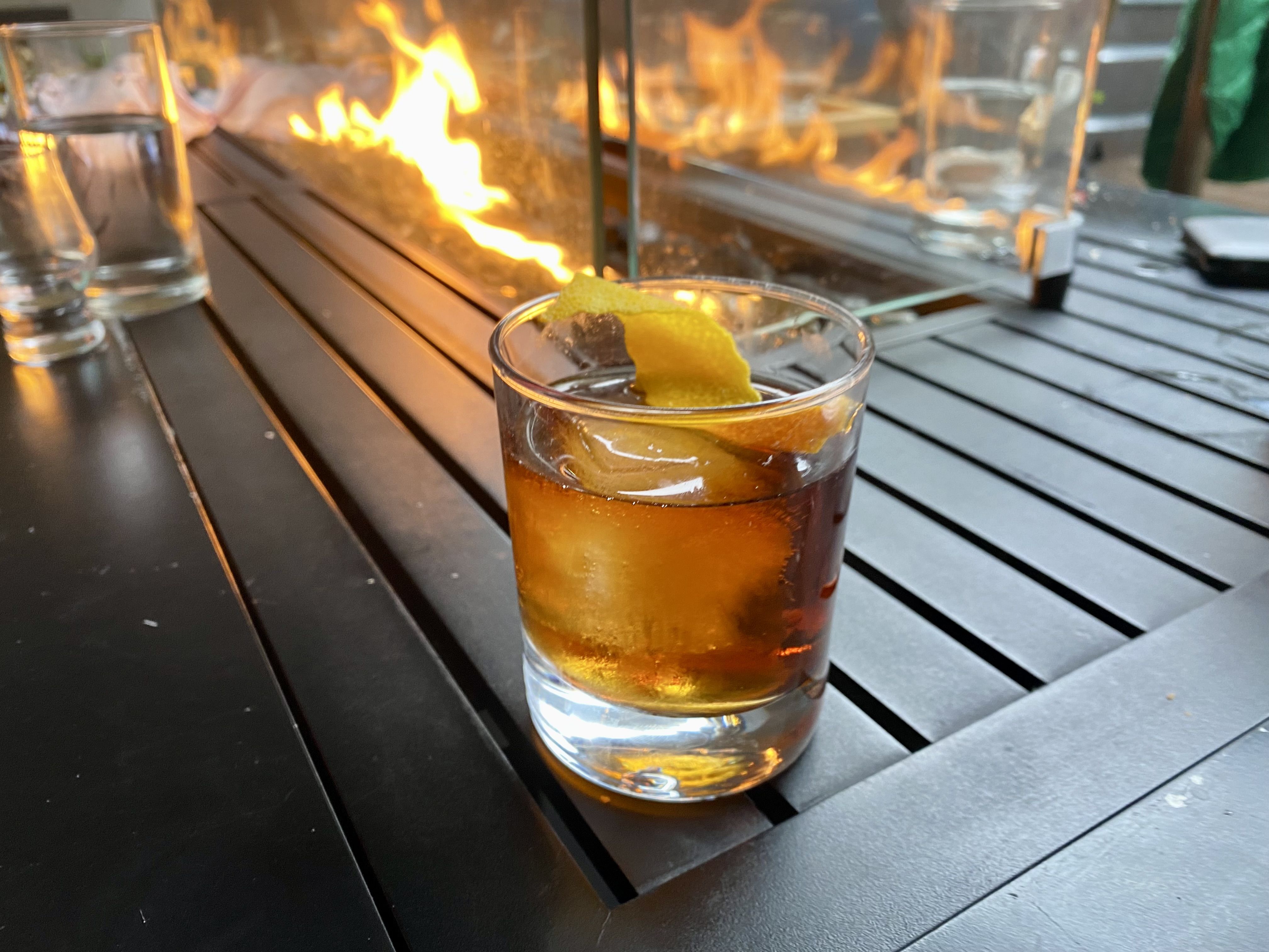 A whiskey drink in a short glass with a twist of lime and a flame in the background.