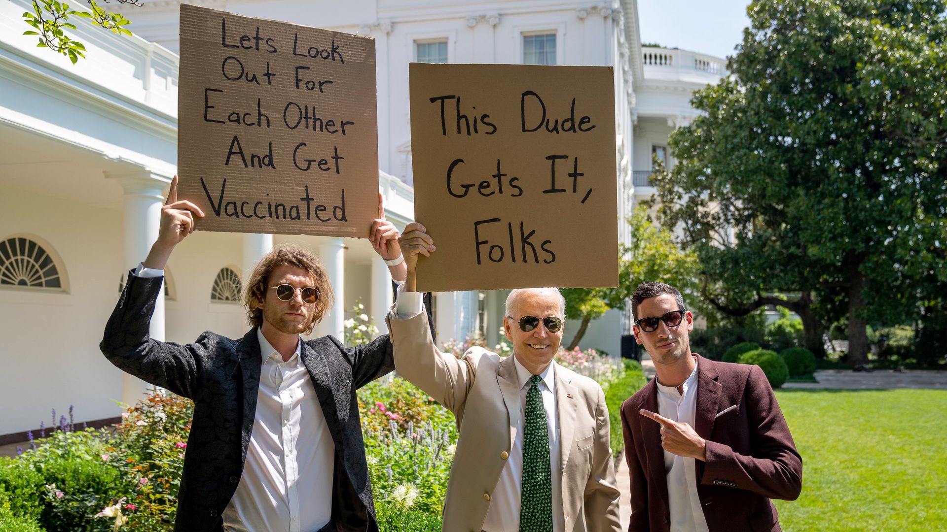 A photo showing President Biden standing between two men, one of whom is holding a sign urging people to get the COVID vaccine.