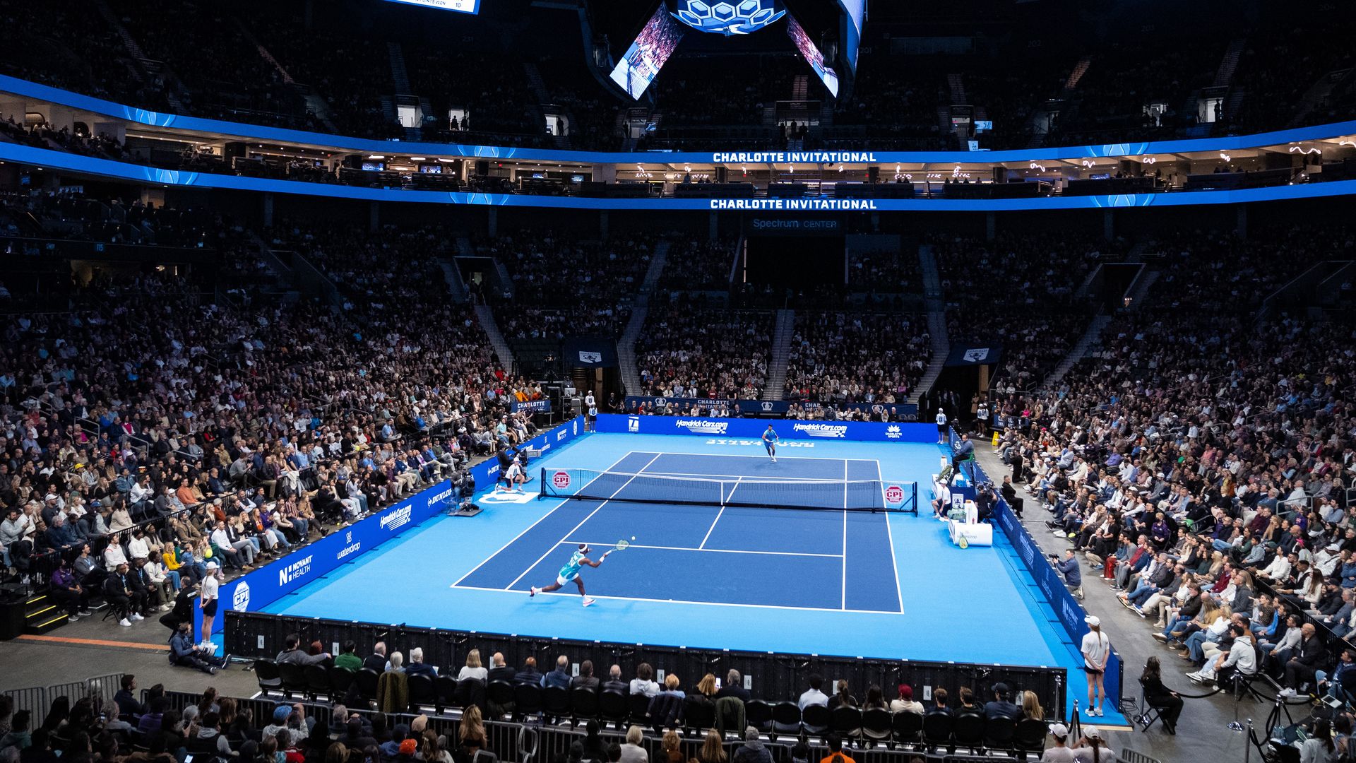 Wide view of indoor tennis match at the Charlotte Invitational, blue courts, packed crowd, players mid-game, large screens and banners with event name above the court.