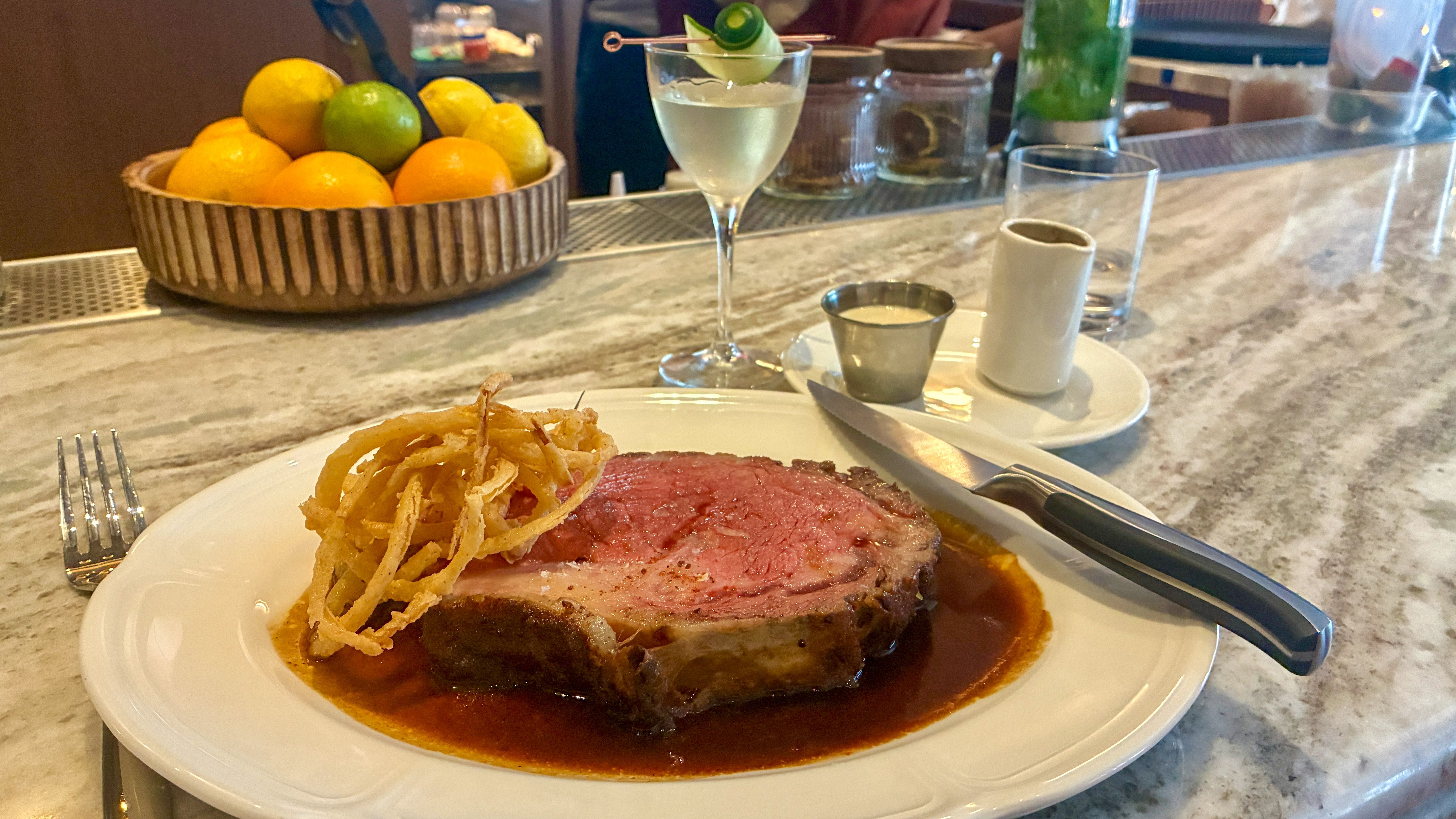 Plate of beef with brown sauce and crispy onion strings on a marble bar; in the background a bowl of lemons and limes, a pale cocktail with cucumber garnish, and glassware.