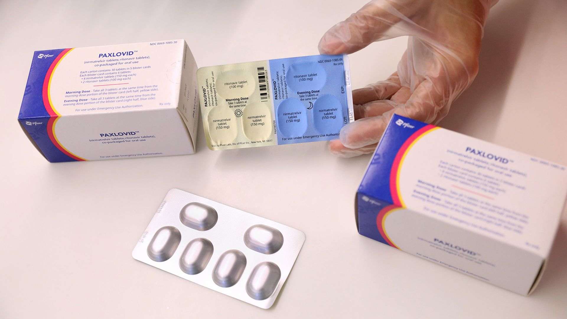 Someone with clear gloves holds out the package of Paxlovid tablets next to two Paxlovid boxes.