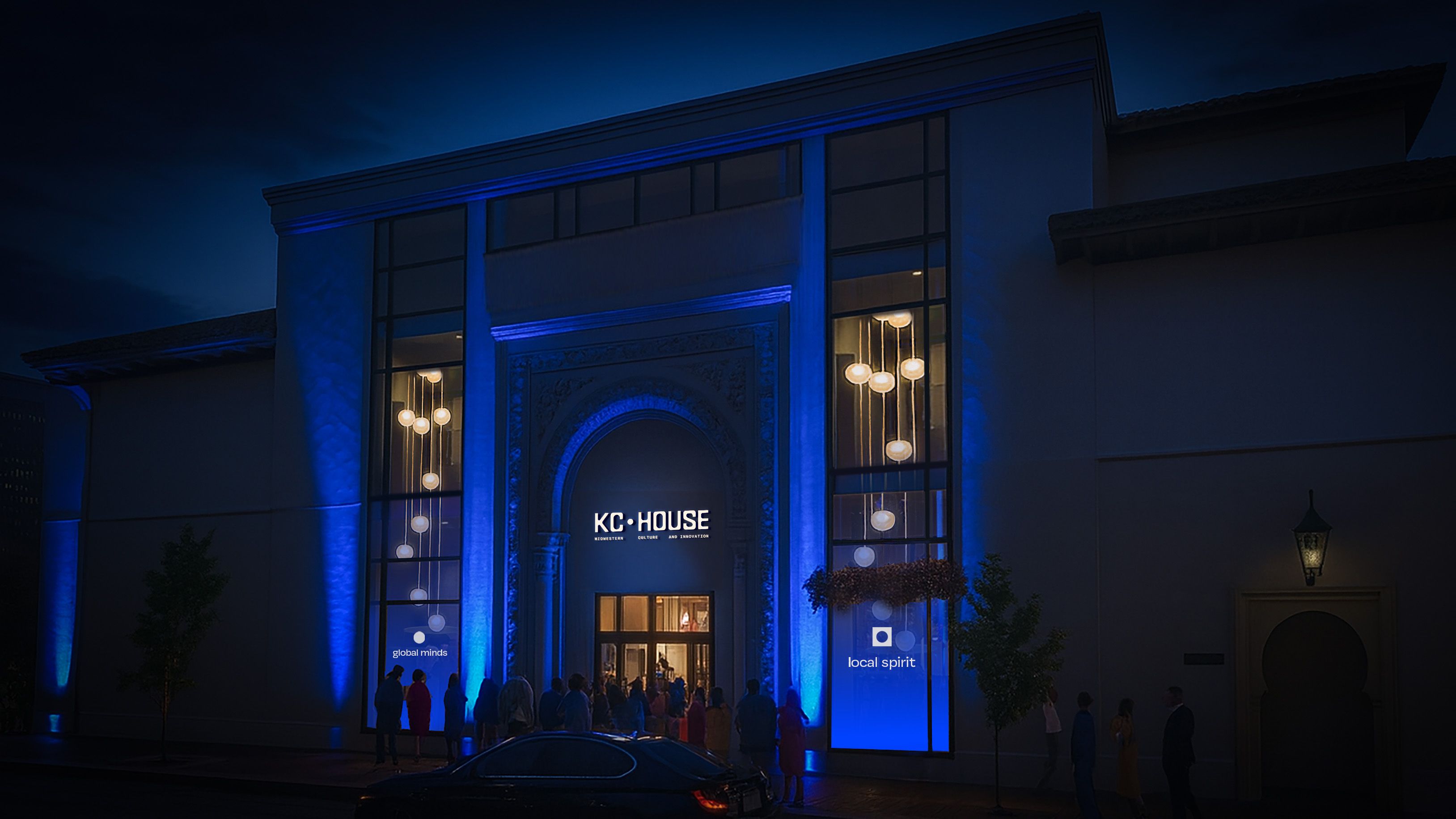 Night view of a modern building with blue lighting, large windows, and hanging lights. People gather outside under a sign reading "KC HOUSE" with phrases "global minds" and "local spirit" visible.