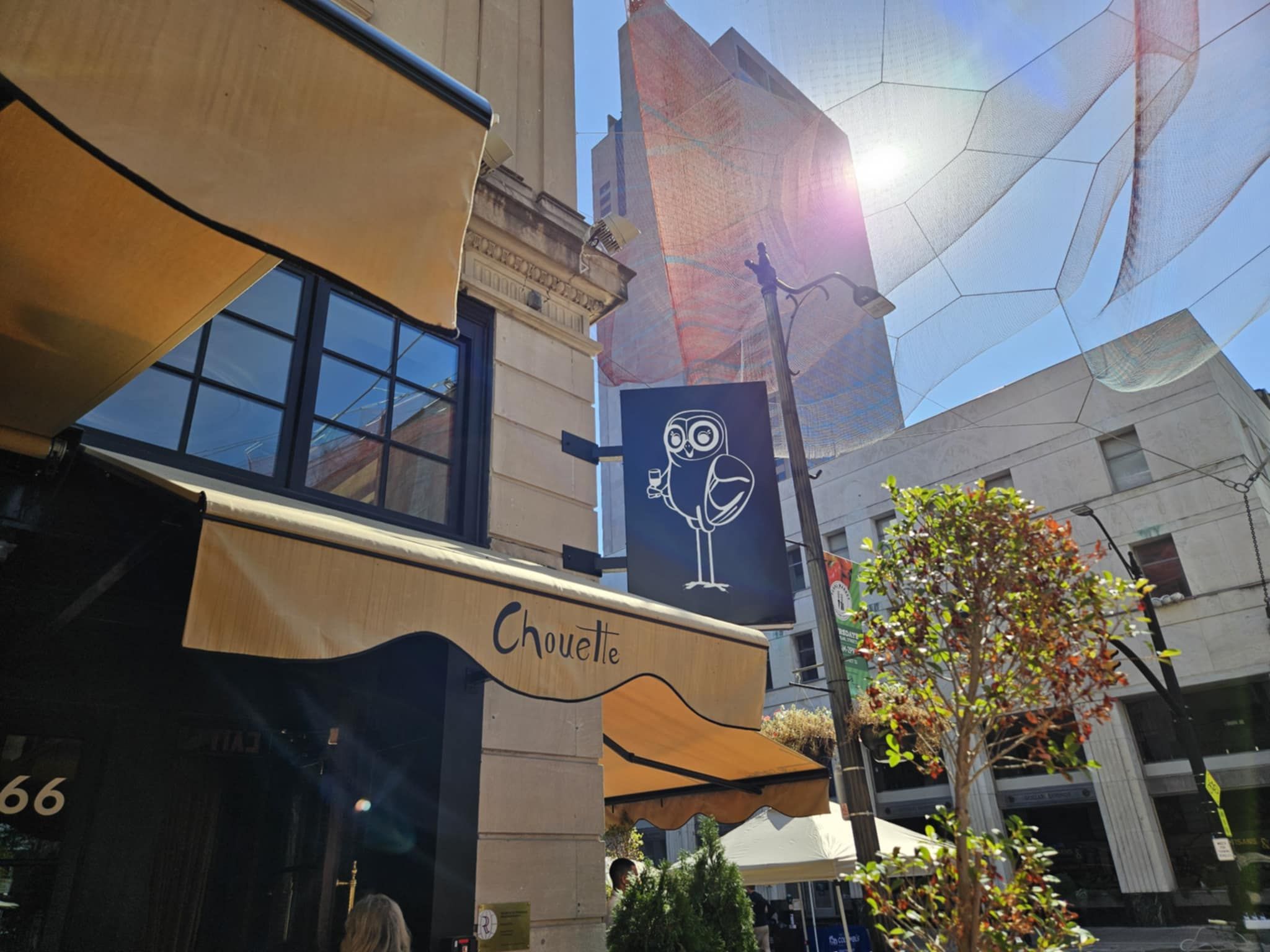 Yellow awning with "Chouette" text outside a building, cartoon owl holding a glass on a blue sign on bright sunny day on a city street with trees and tall buildings.