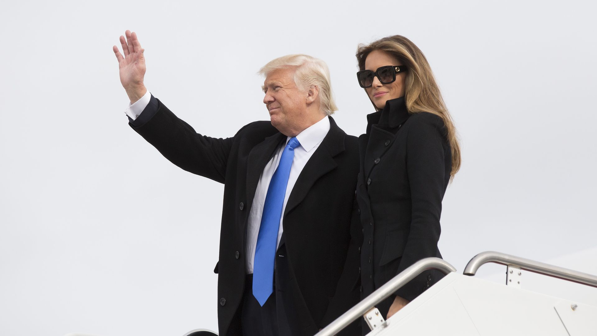 Donald Trump and Melania standing next to each other as the deplane from Air Force One. Trump is waving to a crowd. Melania is wearing all black and sunglasses and is smirking. 