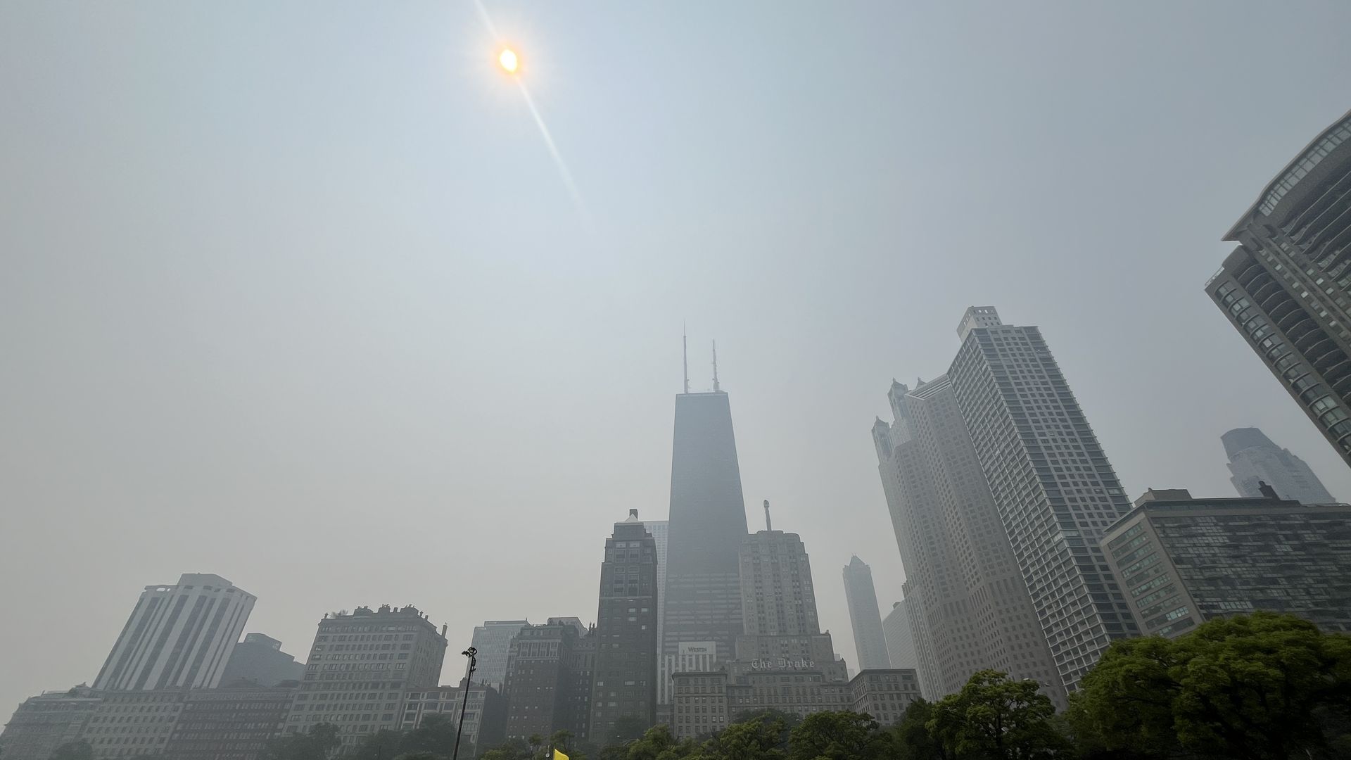 Bright sun and clear sky above a city skyline; a tall dark tapered skyscraper near center, several high-rises to the right, and a sandy foreground with greenery and a railing.