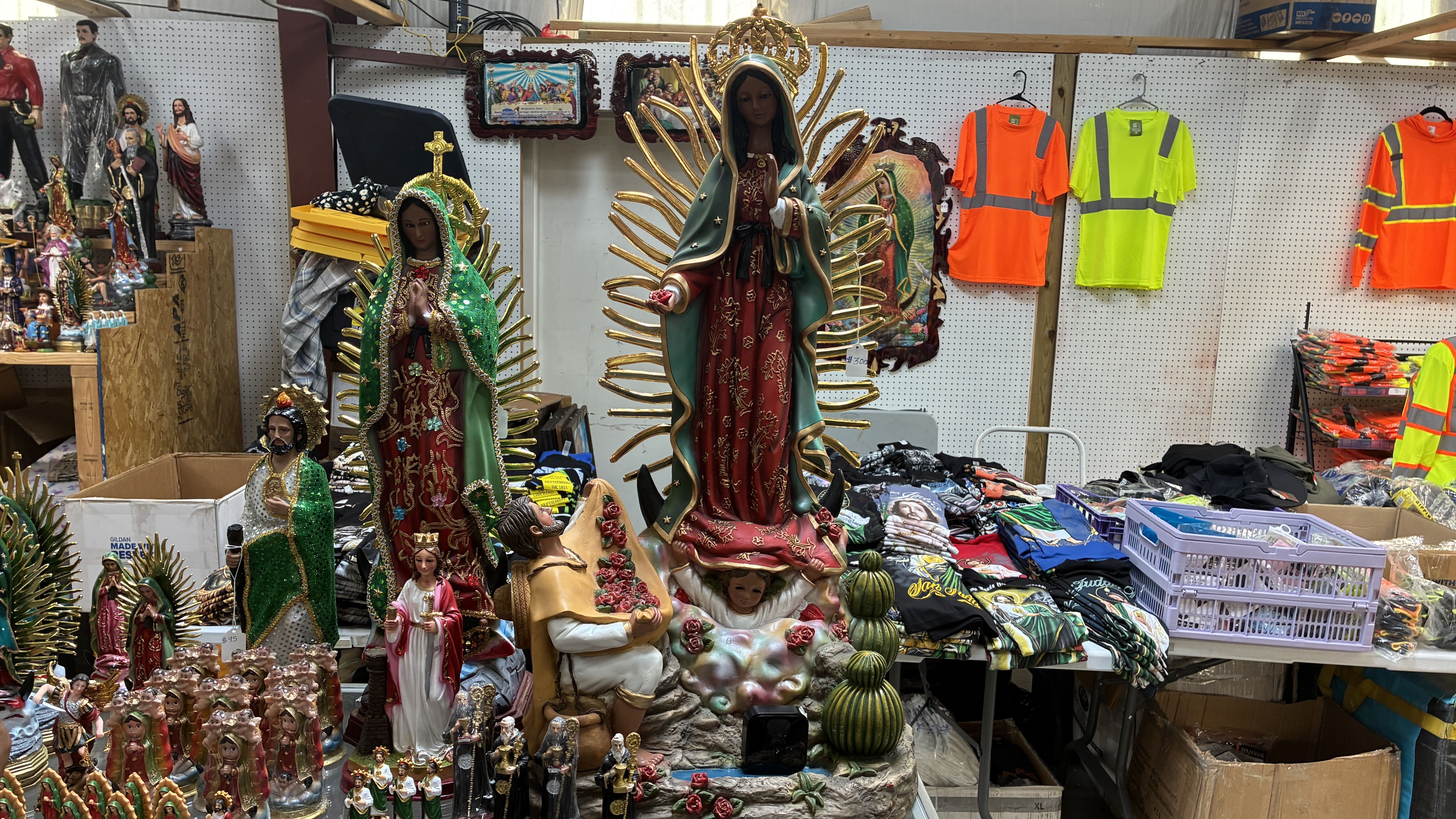 Market stall crowded with religious statues; a large Virgin Mary figure in a green robe with gold rays stands center, surrounded by saints; bright safety vests hang on a pegboard behind.