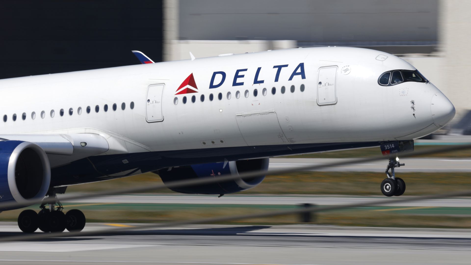 Side view of a white Delta airliner on the runway, cockpit windows at the nose, blue DELTA lettering with a red triangular logo, and extended landing gear, blurred hangar in the background.