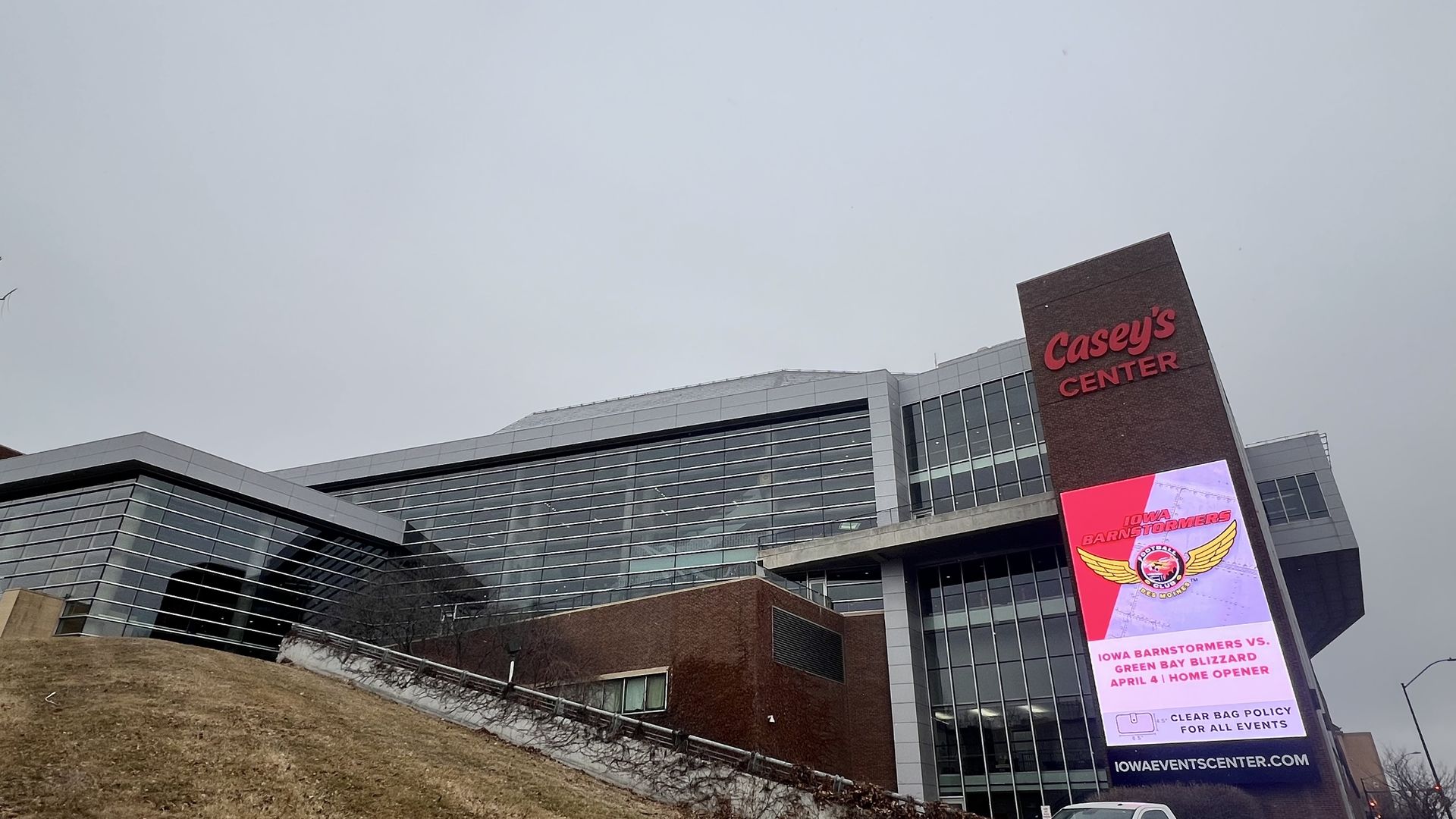Modern building with large glass windows and brick exterior under gray sky, featuring a red sign "Casey's Center" and digital display advertising Iowa Barnstormers vs. Green Bay Blizzard game.