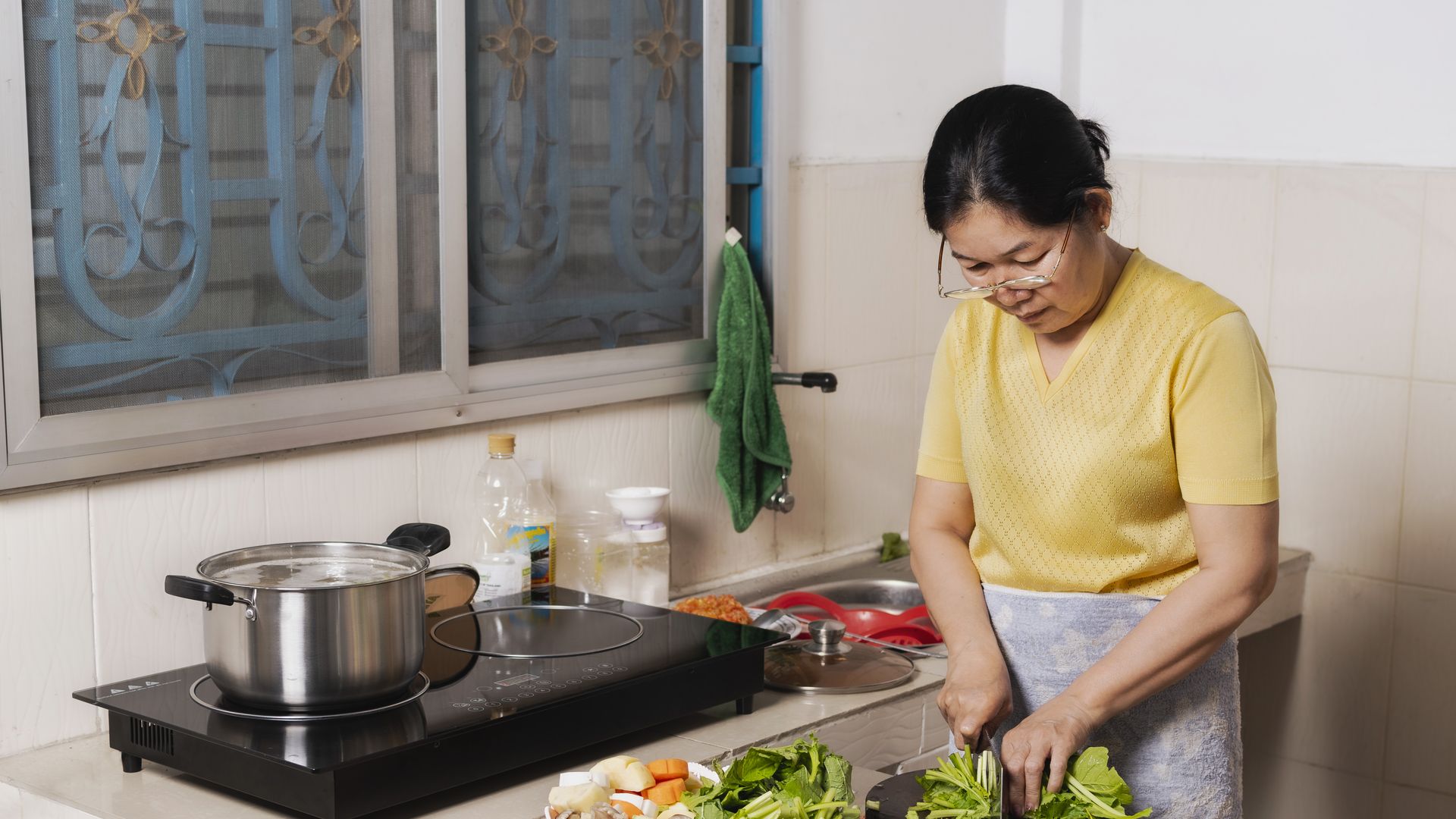 Woman with glasses in a yellow shirt chopping green leafy vegetables in a kitchen with two baskets of assorted veggies and a pot boiling on a black stovetop near a window.