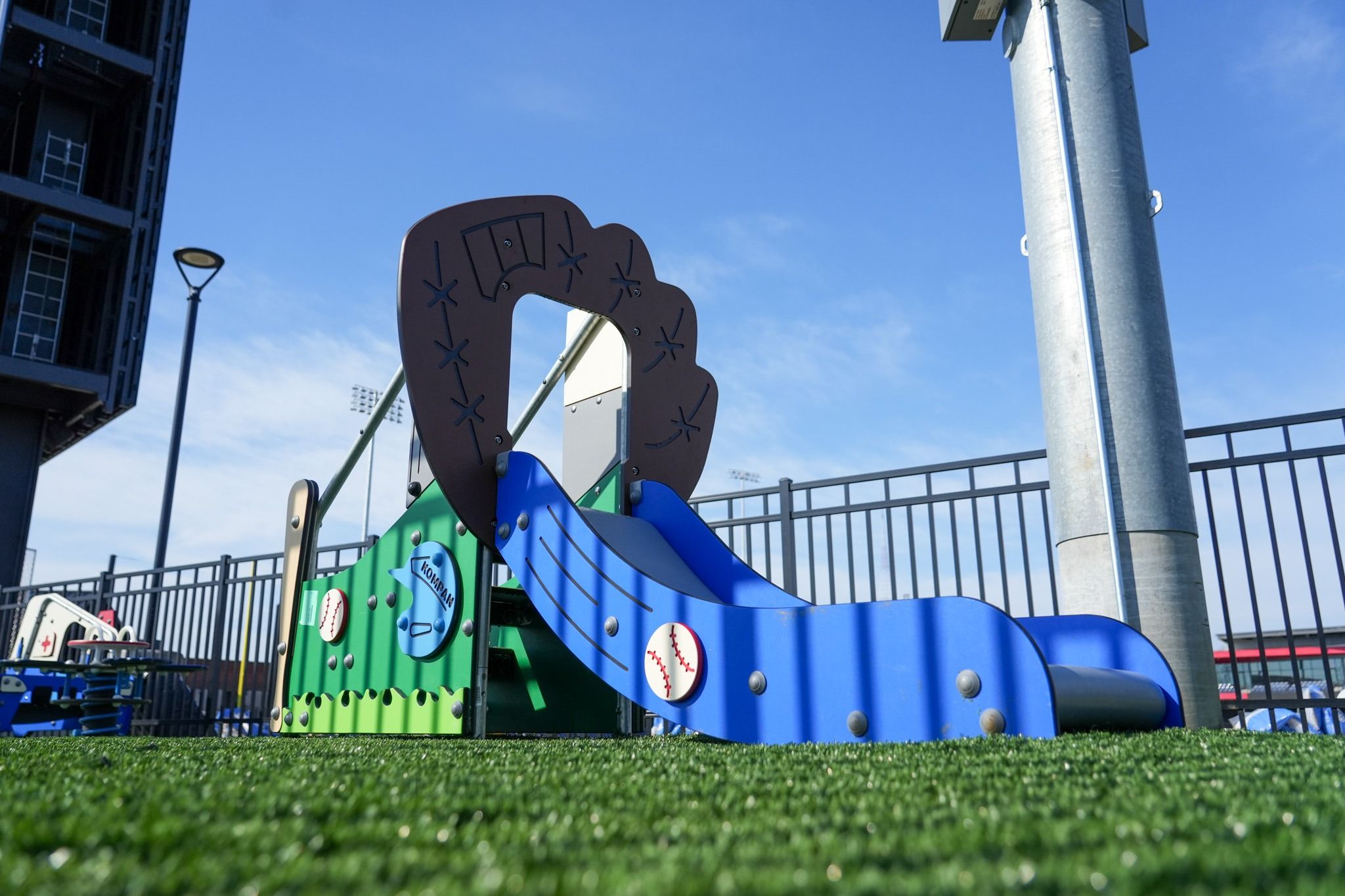Blue playground slide resembling a baseball bat and glove structure on artificial grass with fence and stadium lights in the background on a clear day.