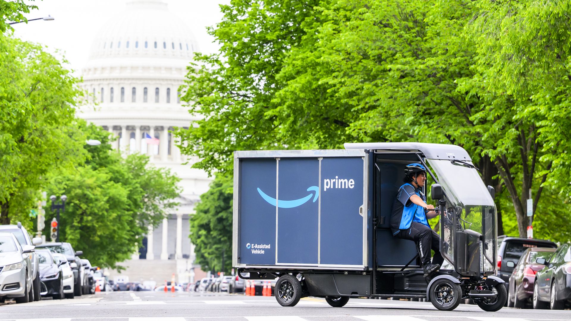 Bright urban street scene with lush green trees framing a distant white U.S. Capitol dome; a blue Amazon Prime delivery bicycle is visible in the foreground