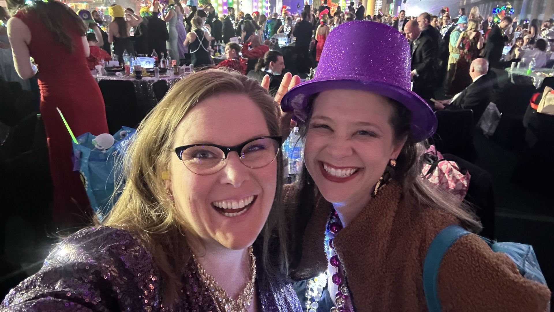Two women smile at a party; one wearing glasses and a purple sequin top, the other with a purple glittery hat and brown jacket. Background shows a crowded festive event with colorful lights.