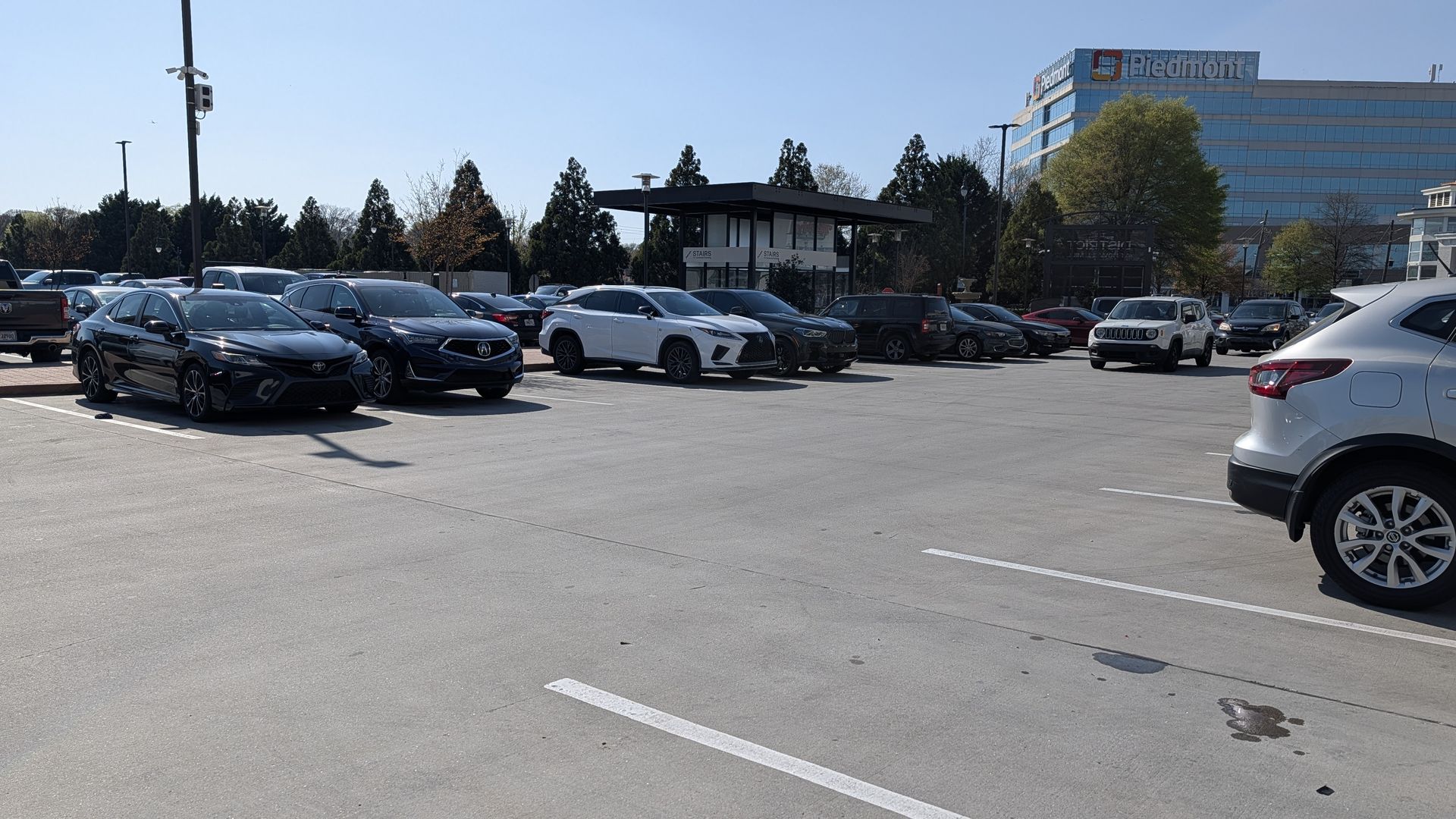 Sunlit outdoor parking lot filled with cars: black, white, and silver SUVs and sedans, with a glass office building featuring a large PIEDMONT sign and a modern glass pavilion in the center.
