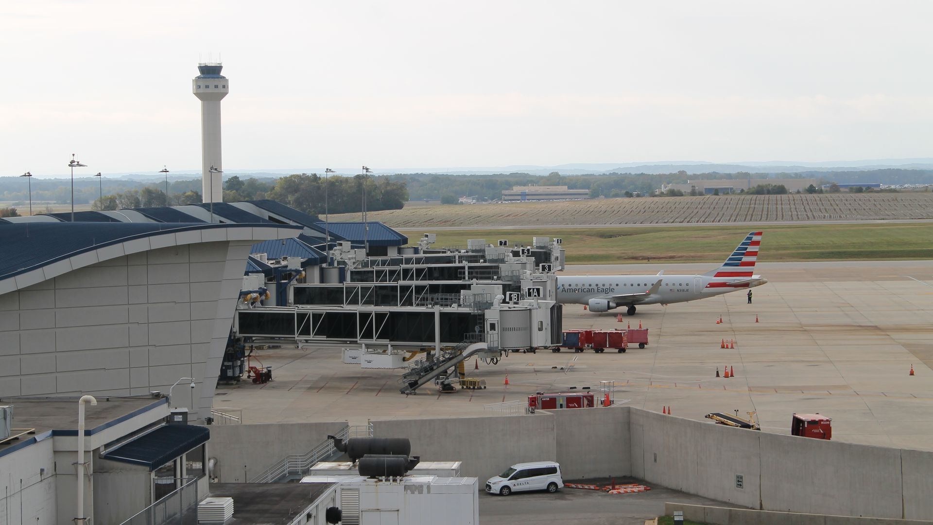 View of an airport terminal with empty jet bridges and an American Eagle airplane on the tarmac, ground vehicles, control tower, and distant landscape under a cloudy sky.