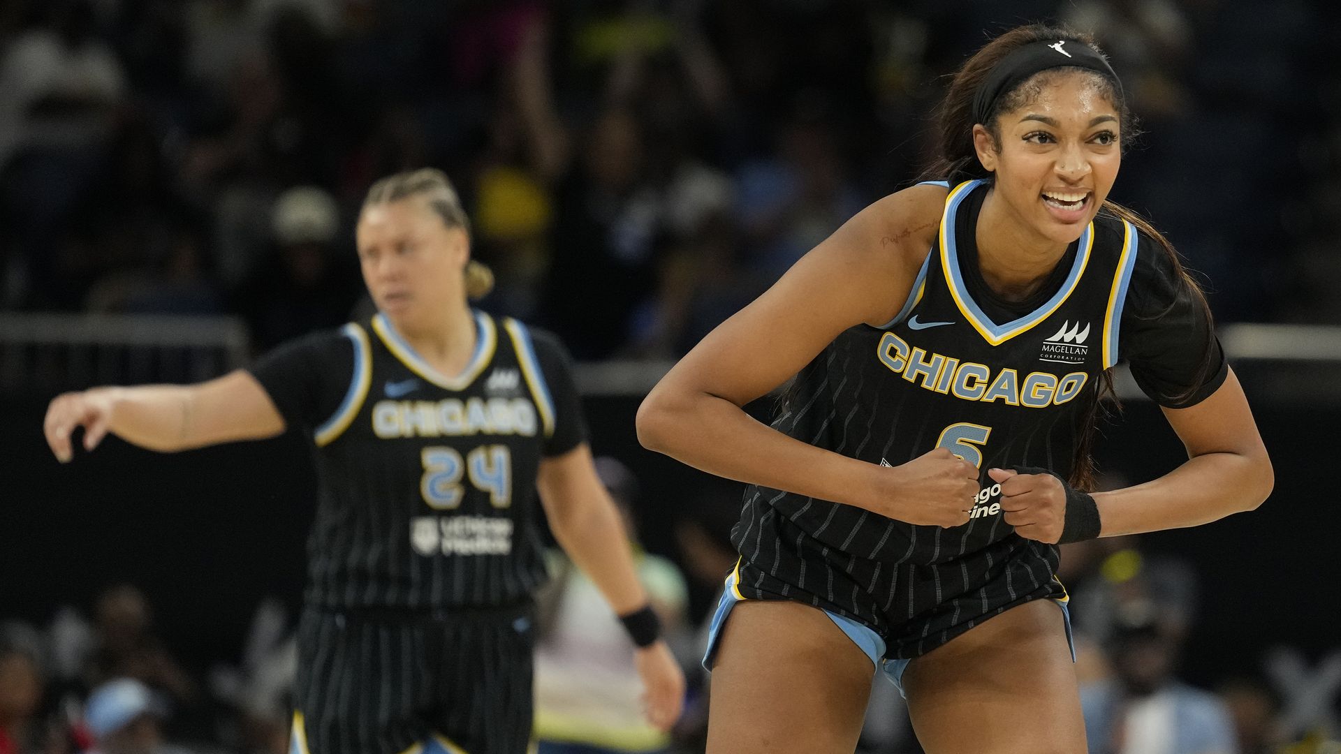 Two Chicago Sky players in black pinstriped jerseys with blue and yellow trim; foreground player #5 smiles and runs, while teammate #24 stands in the background on a basketball court.