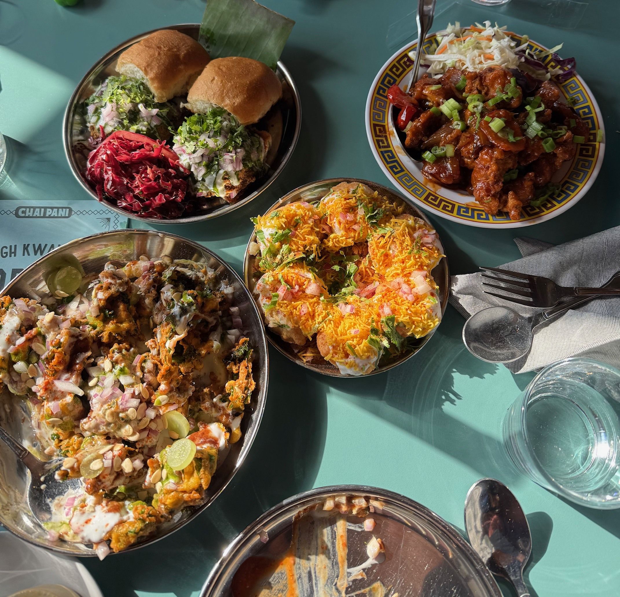 Top-down view of a teal table featuring assorted dishes: two mini burgers with greens, a plate of saucy fried chicken bites with scallions, a bowl of colorful chaat with yogurt and onions, and a cheesy, topped dish.