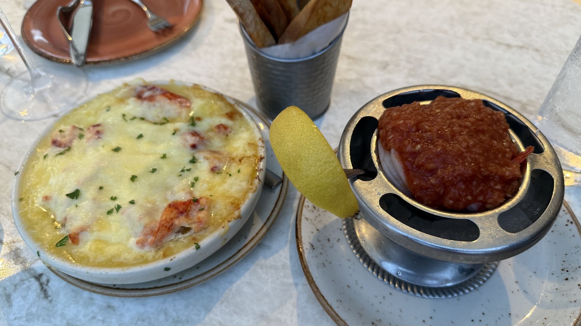 White ramekin dish filled with melted cheese over pieces of lobster on a marble surface; to the right, a metal ring holds a red sauce and shrimp dish with a lemon wedge. A cup of crusty bread slices sits nearby.