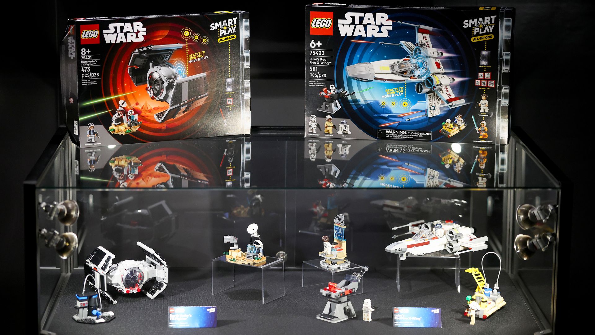 Two LEGO Star Wars boxed sets in a glass display case: left box with red/black artwork, right box with blue artwork and an X-wing; several small LEGO builds and minifigures on the shelf.