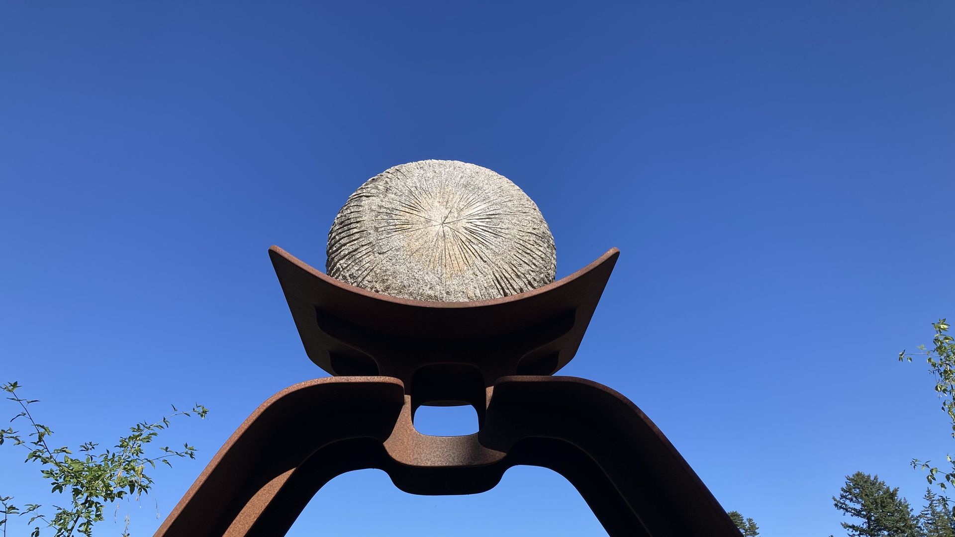 A rust-colored three-legged metal sculpture supports a large round stone against a bright blue sky, with trees in the background.