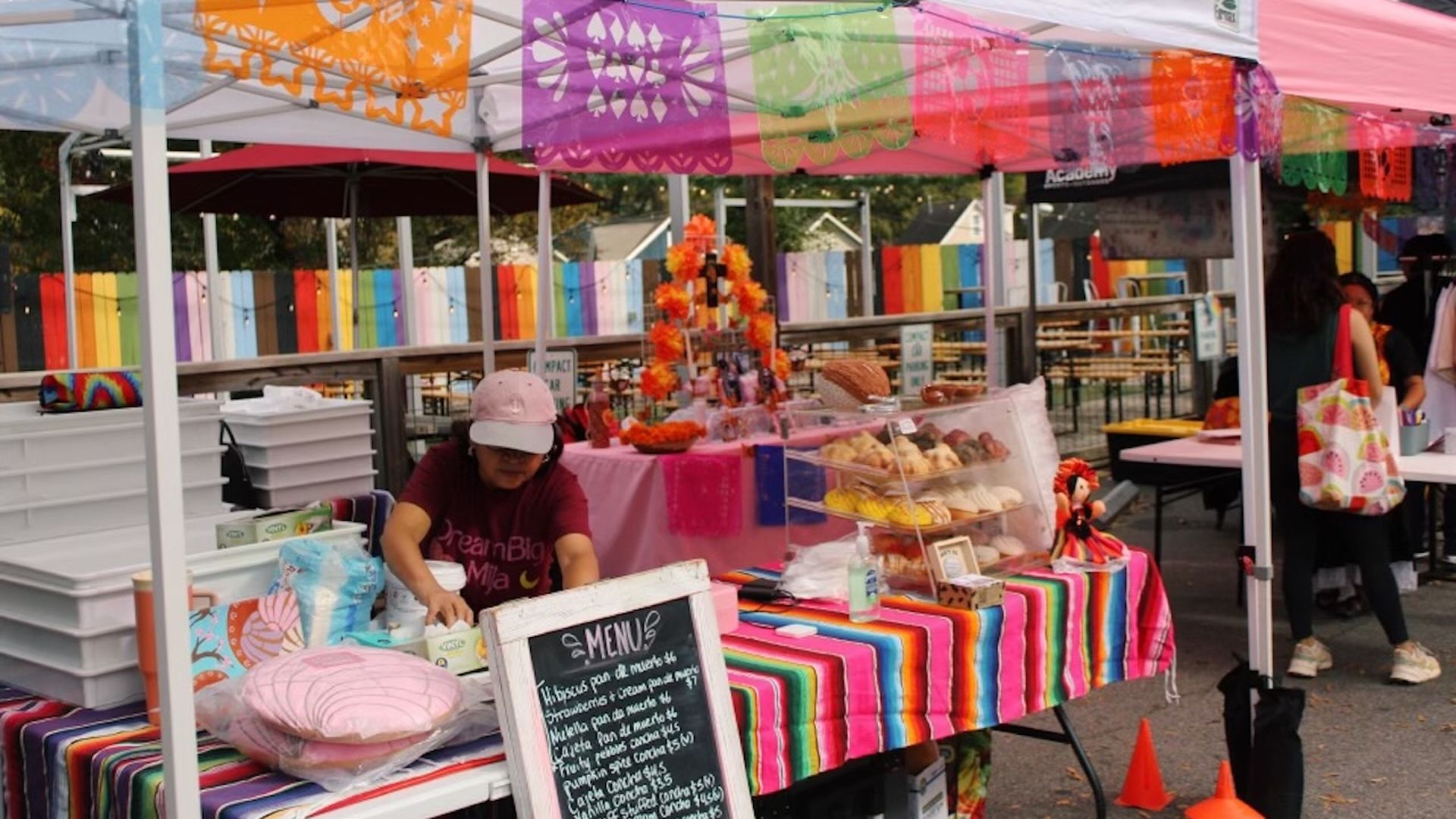 Outdoor food stall with colorful striped tablecloths and papel picado decorations, a vendor wearing a cap arranging items, display case with pastries.