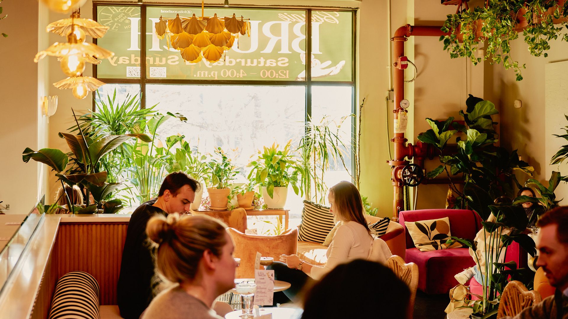 A cozy cafe interior with warm lighting and plants. People are sitting at tables, engaged in conversation. The furniture includes striped seating and colorful chairs. Overhead, stylish light fixtures hang from the ceiling. Large windows at the back fill the space with natural light.